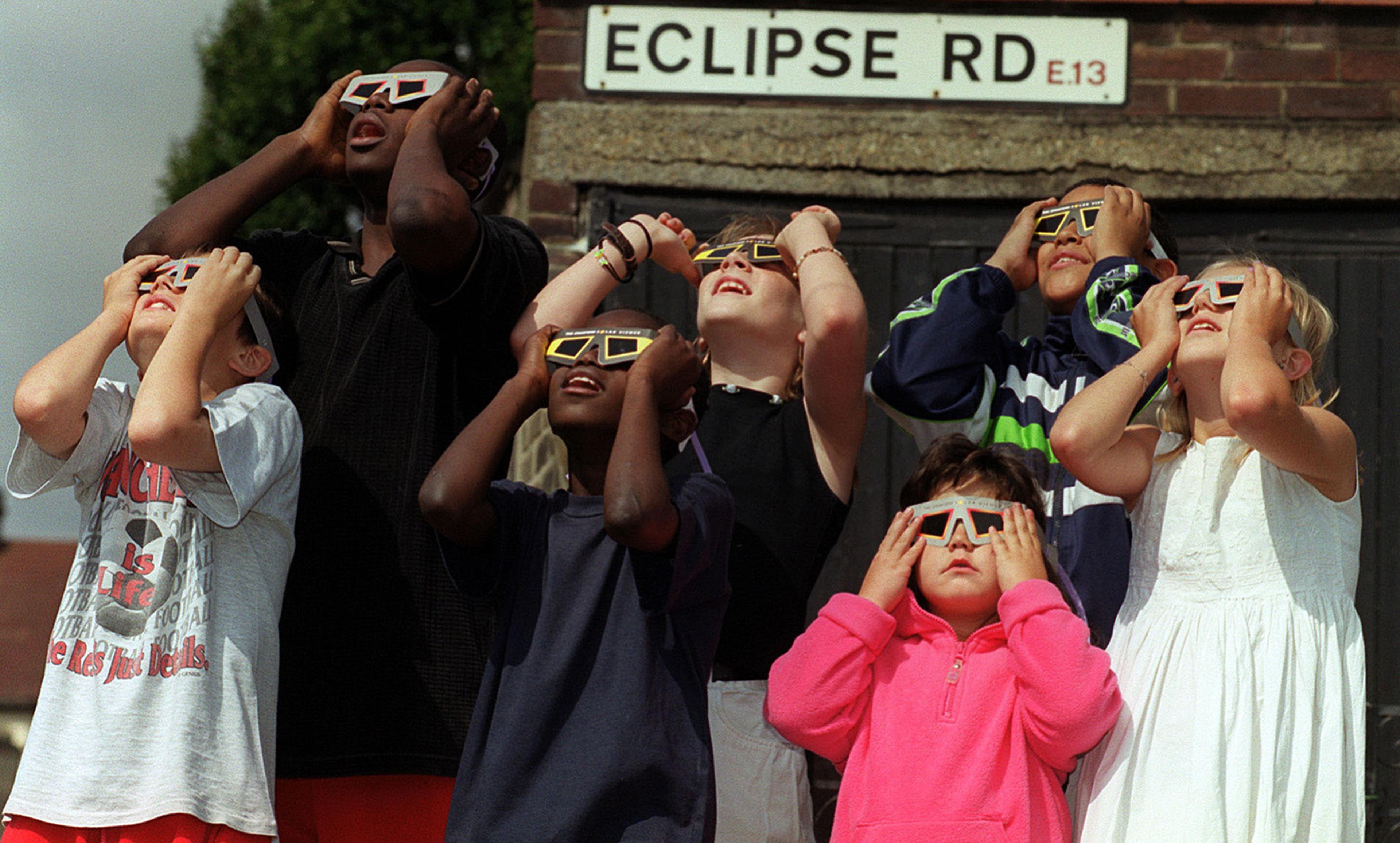 Seven children wearing eclipse glasses looking up at the sky under a street sign reading “ECLIPSE RD”.