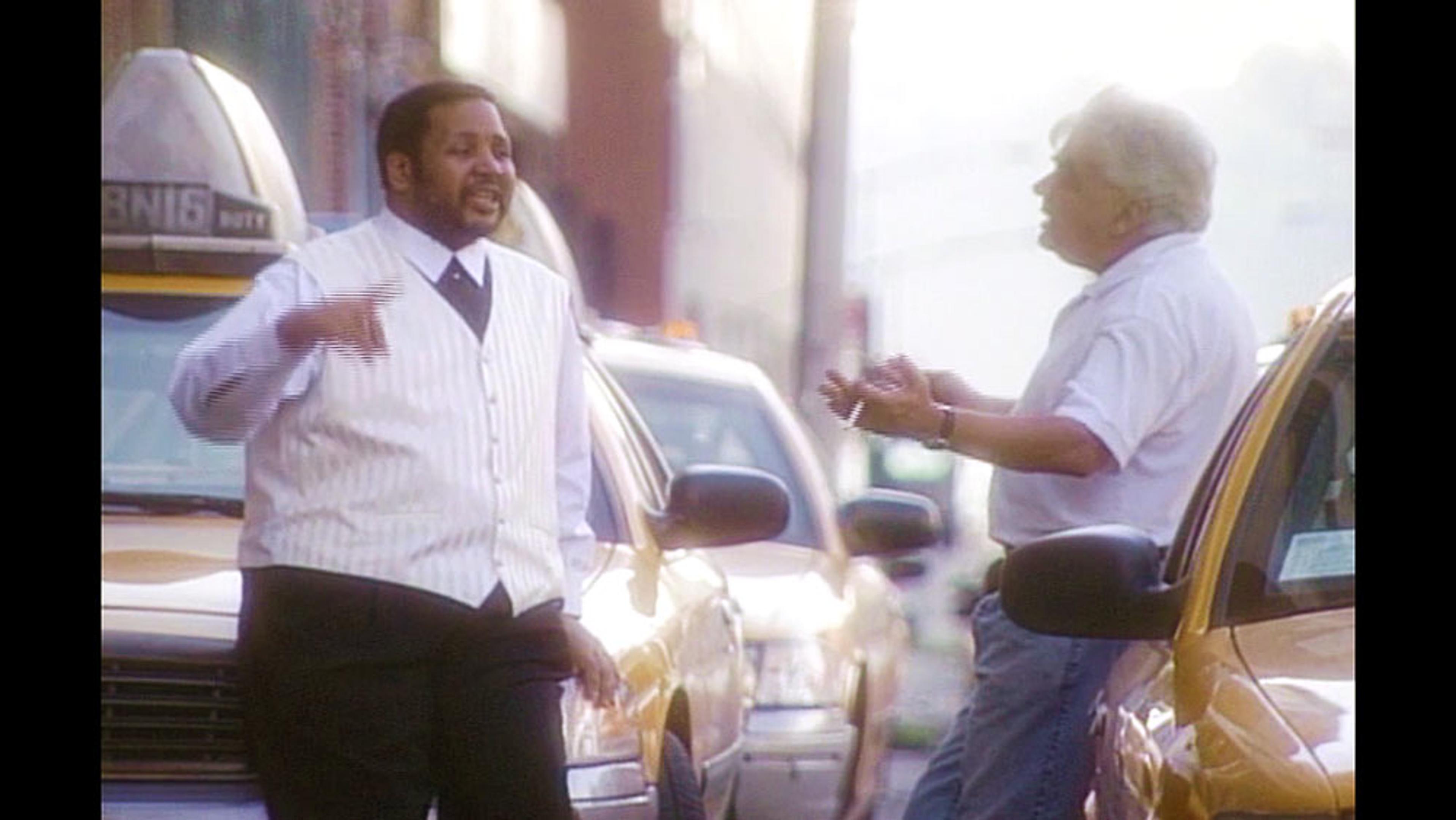 Two men talking on a street near yellow taxis, one wearing a white shirt and waistcoat, the other in a short-sleeve shirt.
