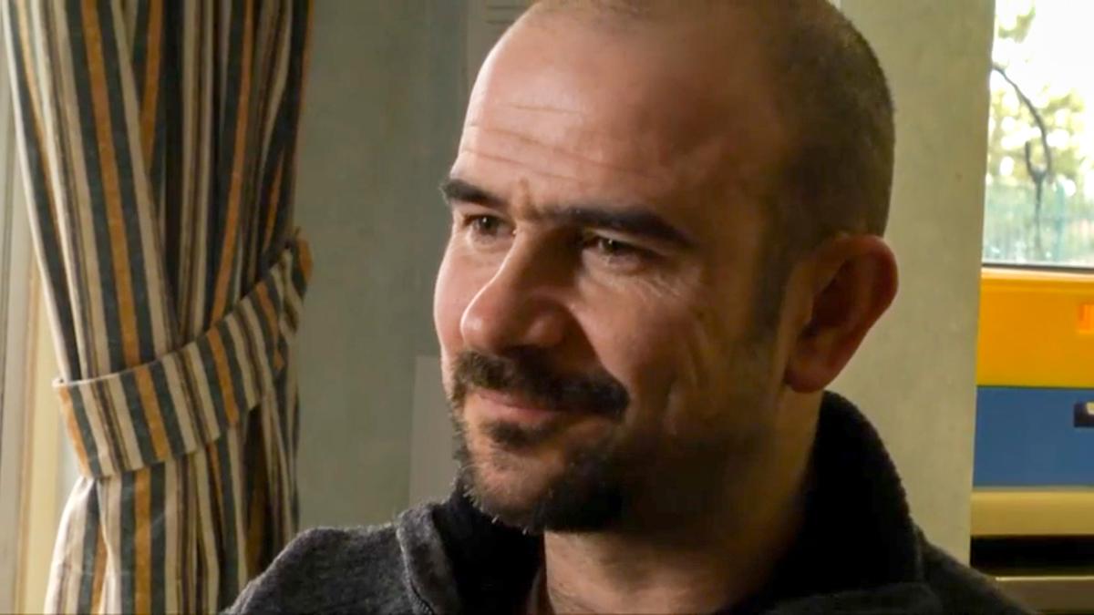 Photo of a bald man with a beard smiling, indoors with striped curtains in the background.