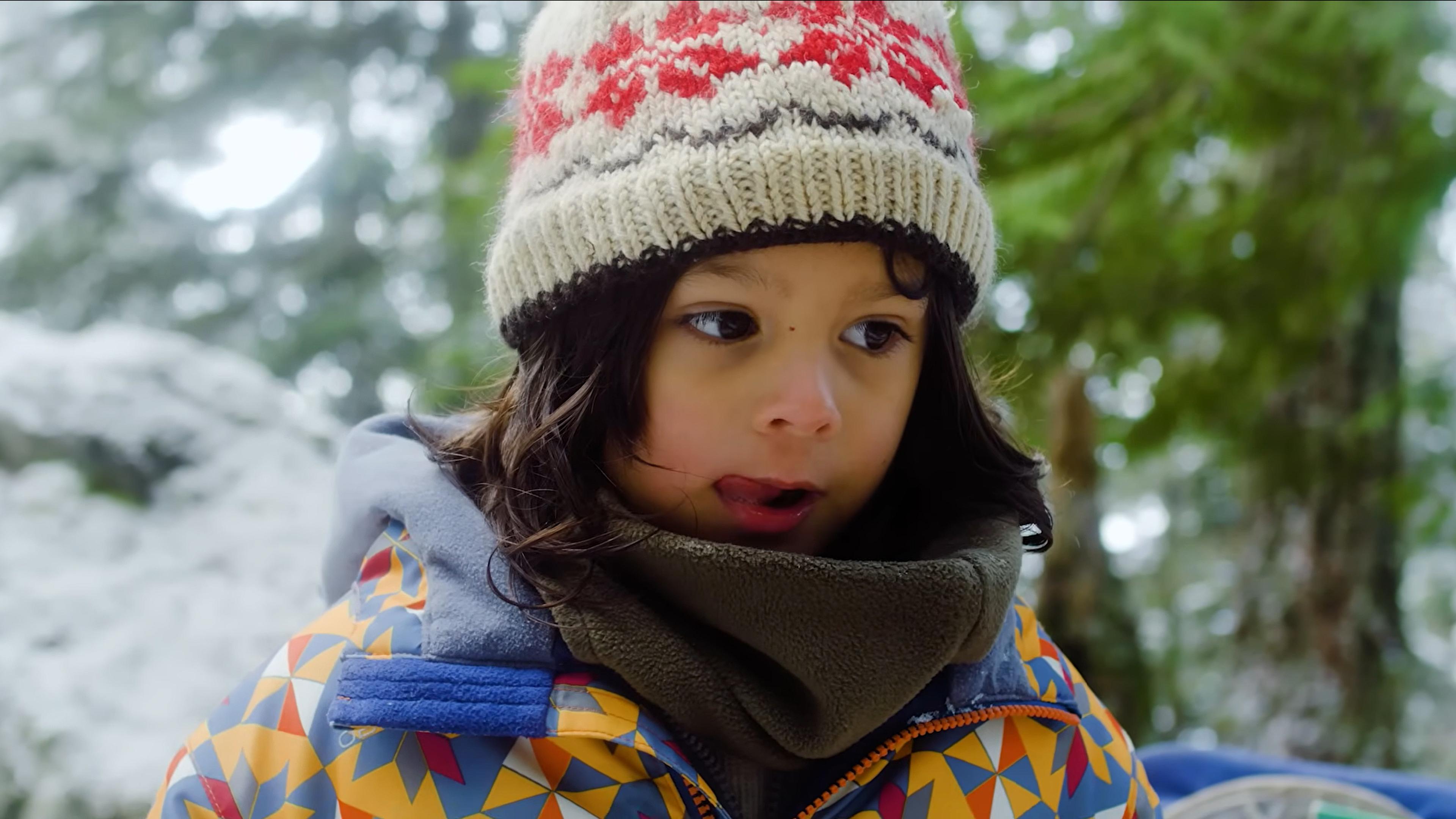 Photo of a child in a colourful winter jacket and knitted hat, in a snowy forest, with a thoughtful expression.