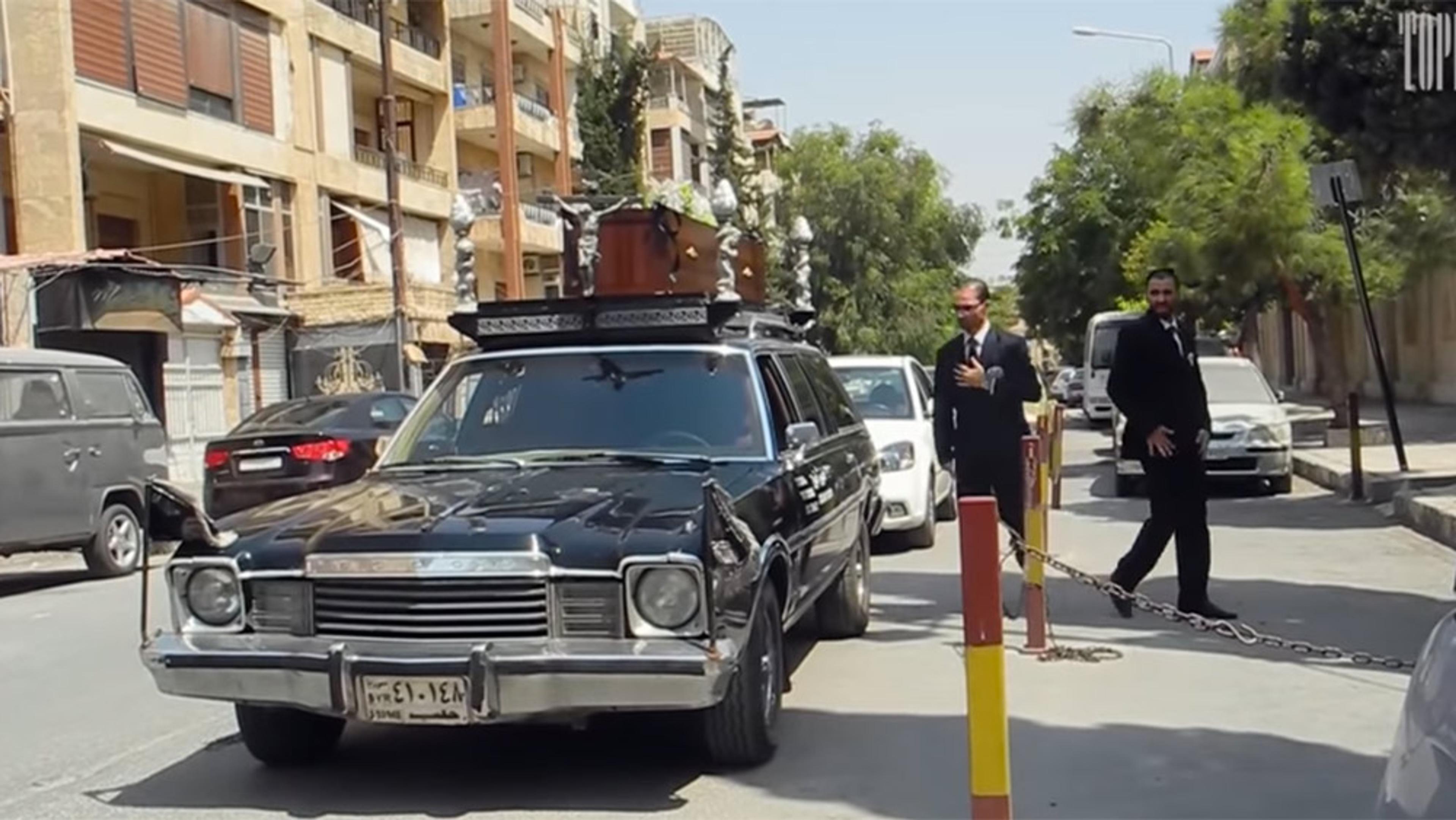 A black hearse with ornate coffin on the roof, parked on a street with two men in suits standing nearby.