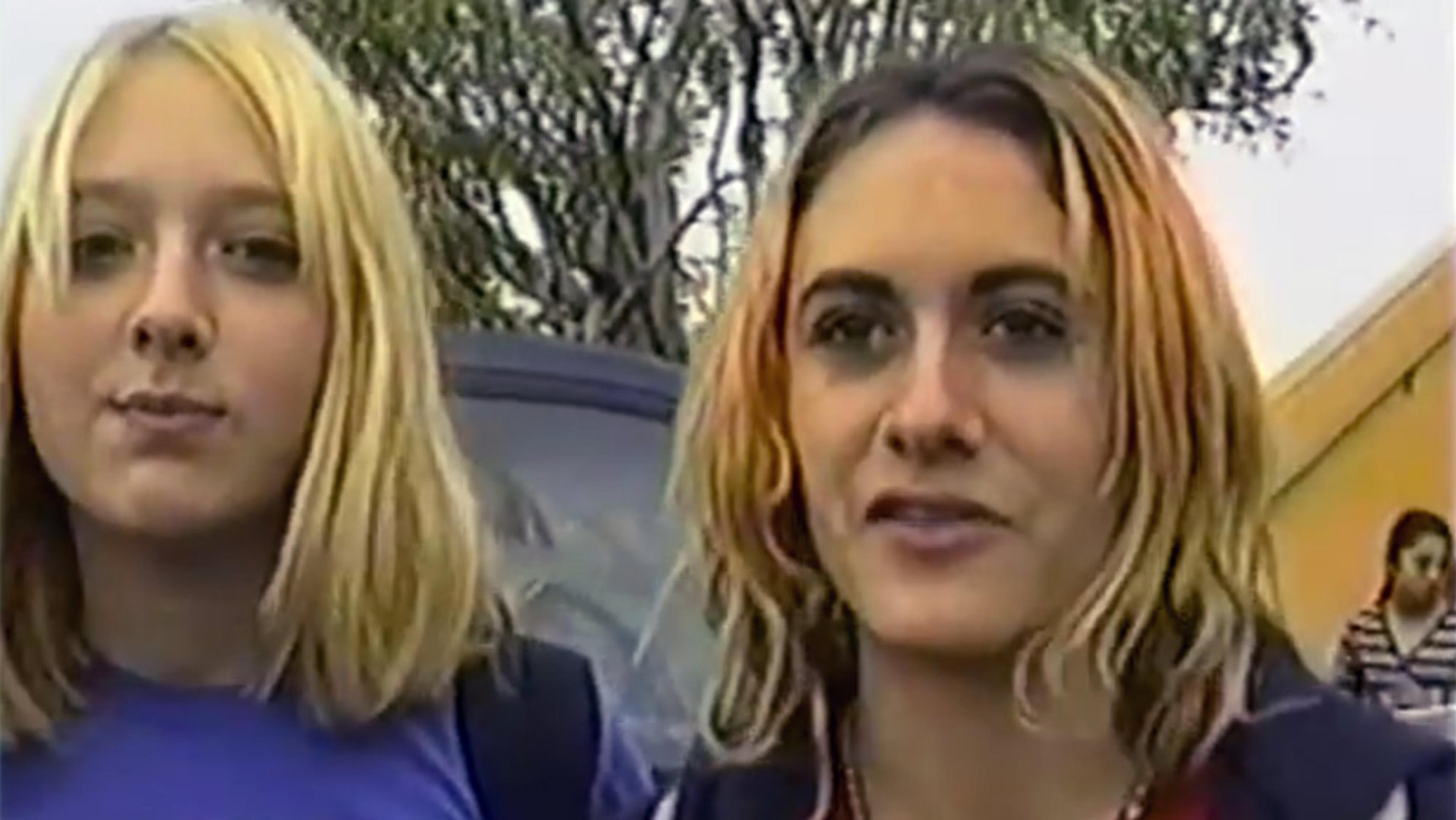Grainy 90s-looking video still of two young women with dyed blonde hair in an outdoor setting, standing in front of a car.