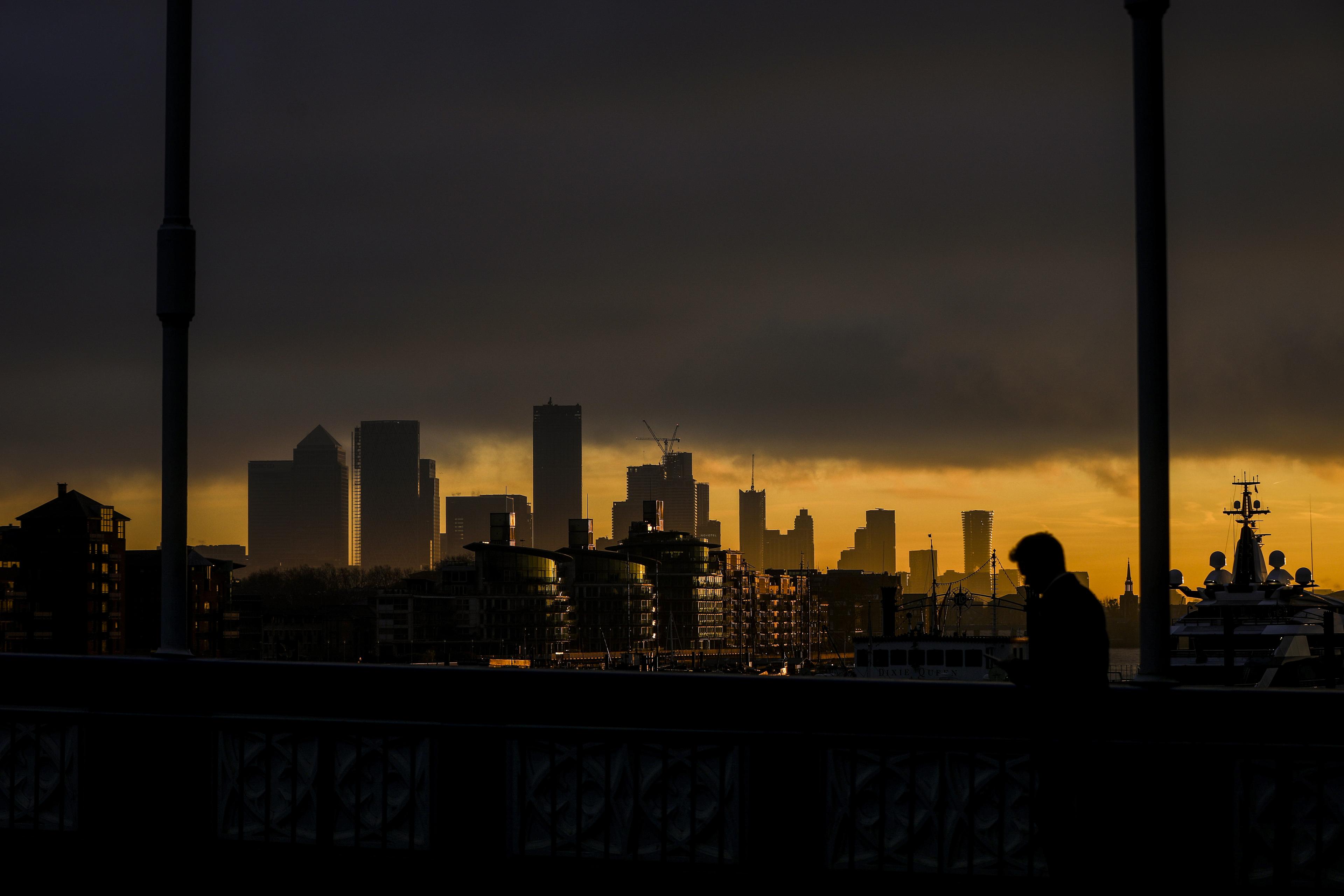 Photo of a cityscape at sunset with silhouetted buildings and a person on a bridge in the foreground.