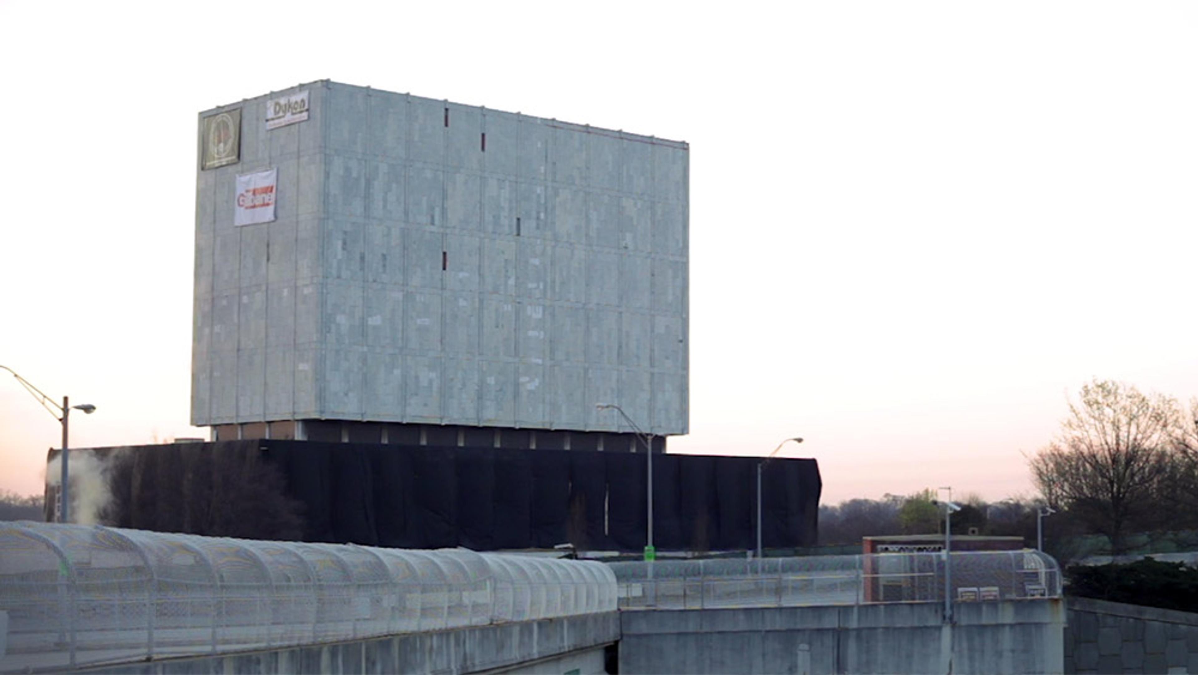 A large rectangular concrete building with minimal windows, partially wrapped in black material, against a sunset sky.