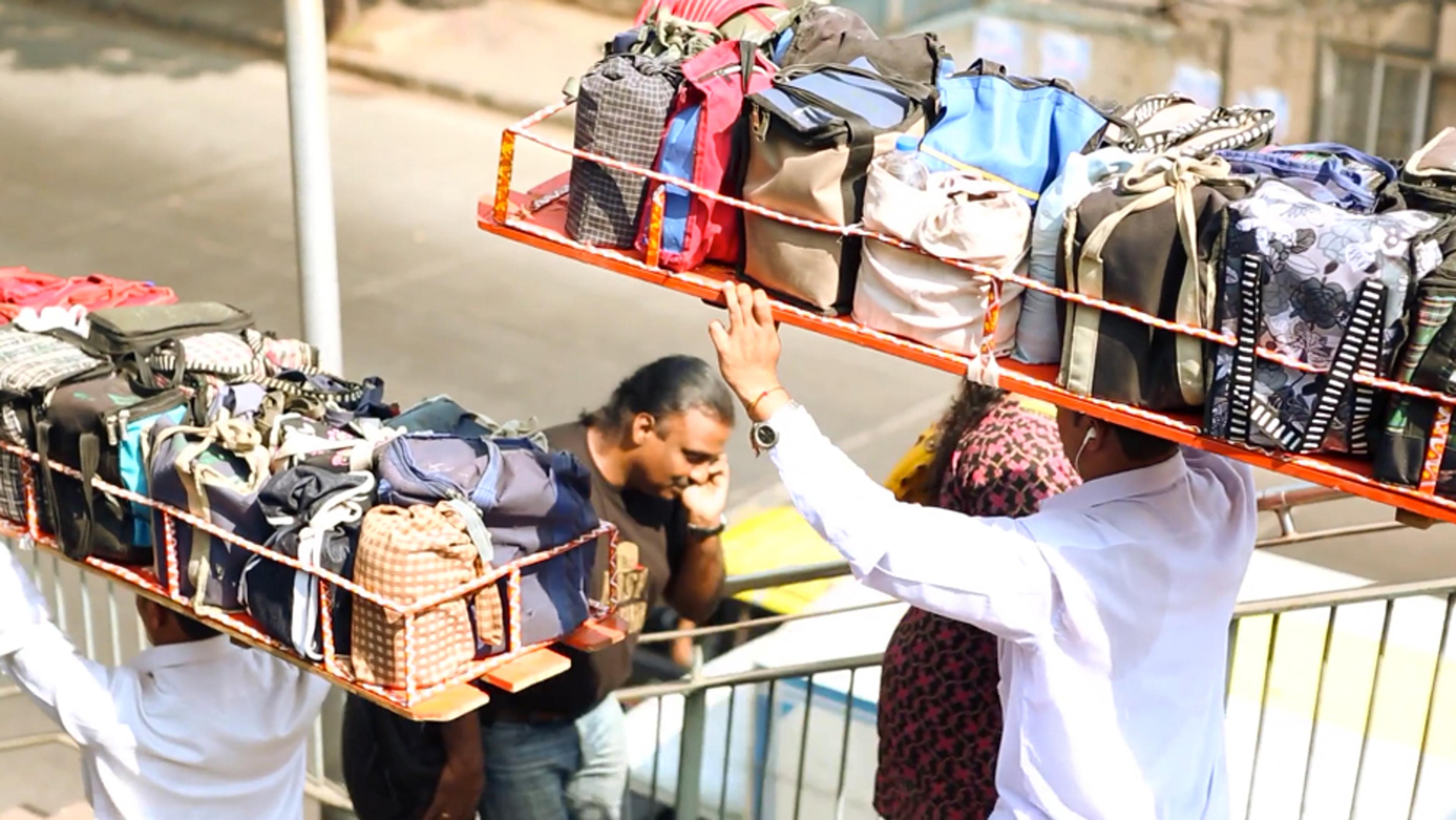 Two people carrying trays of colourful bags on their heads while walking on a street with another person using a phone.