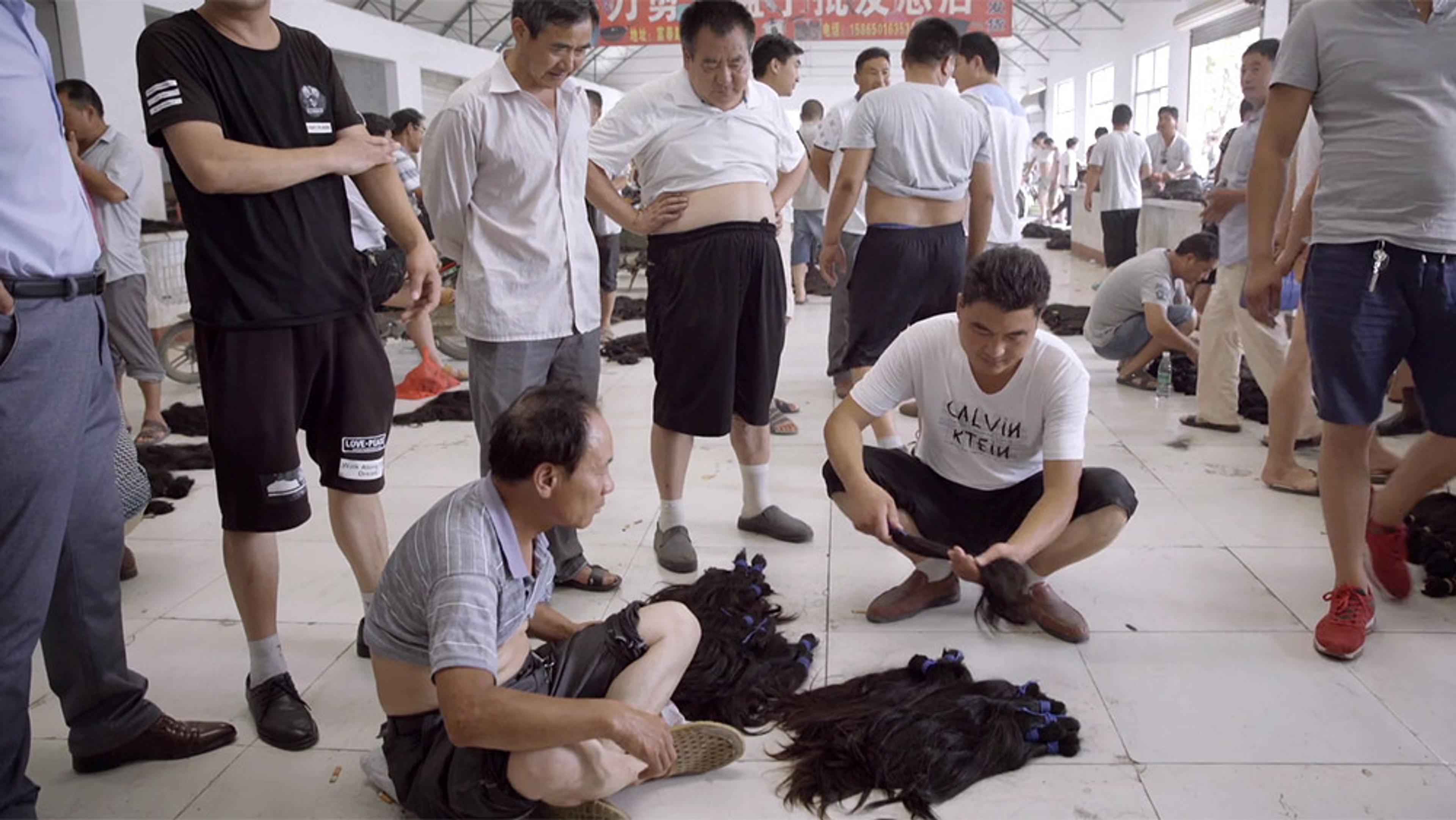 A busy indoor market where men inspect and handle bundles of black hair on the floor, with people standing and squatting nearby.