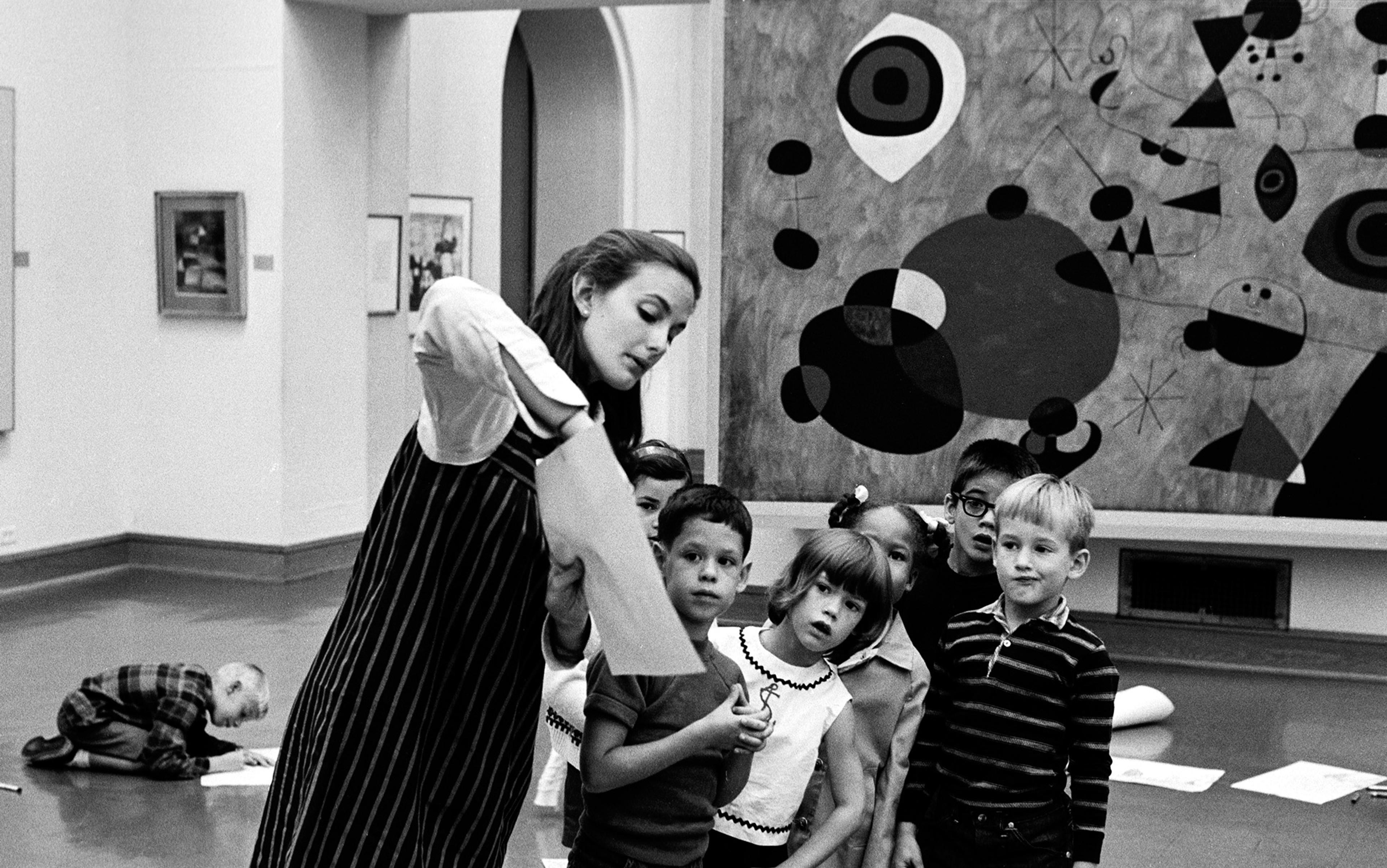 Period photo of a woman showing something on paper to a diverse group of children in an art gallery. Abstract paintings are visible in the background.