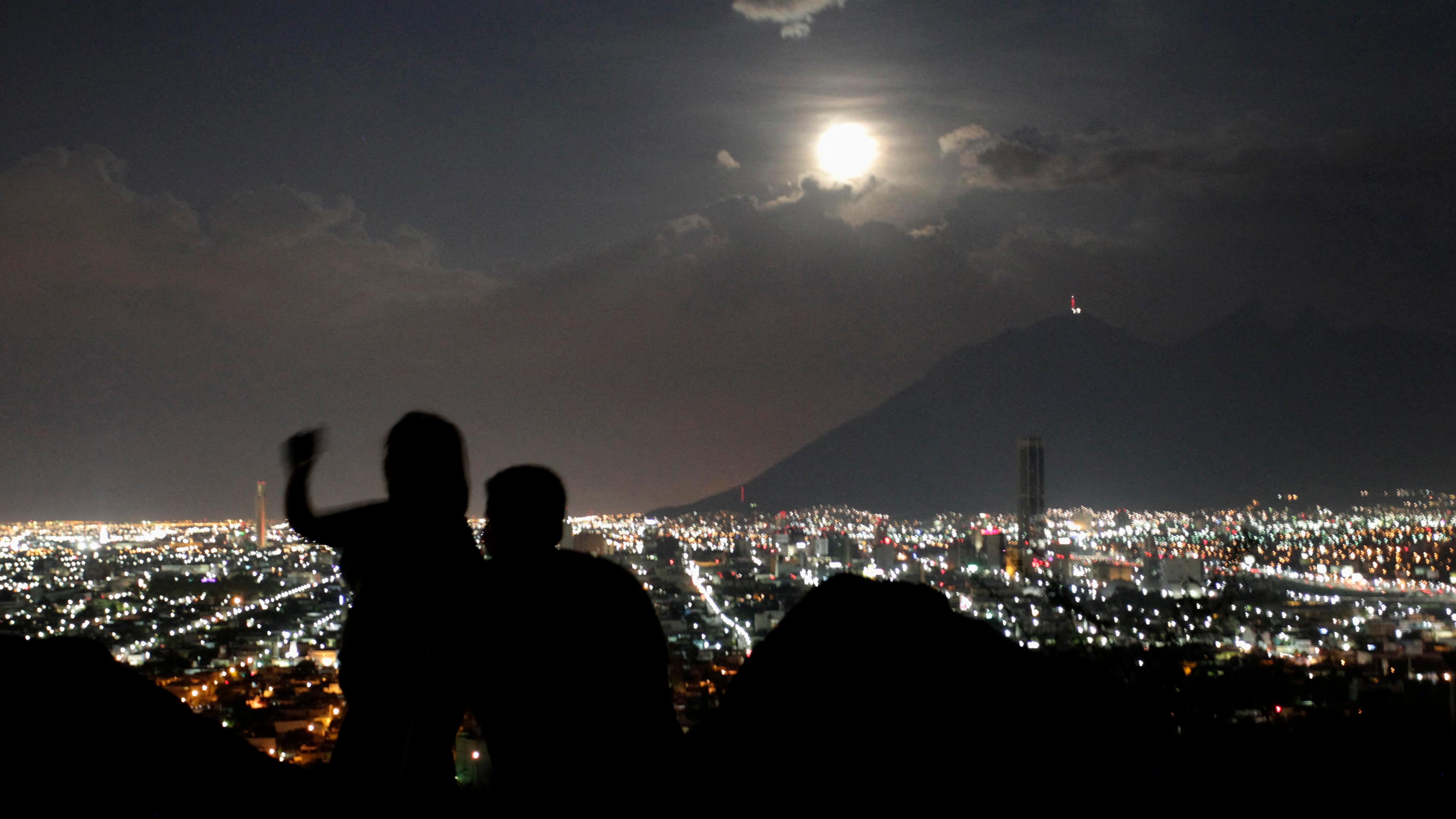 A cityscape at night with two silhouetted figures in the foreground and a full moon above a mountain in the background.