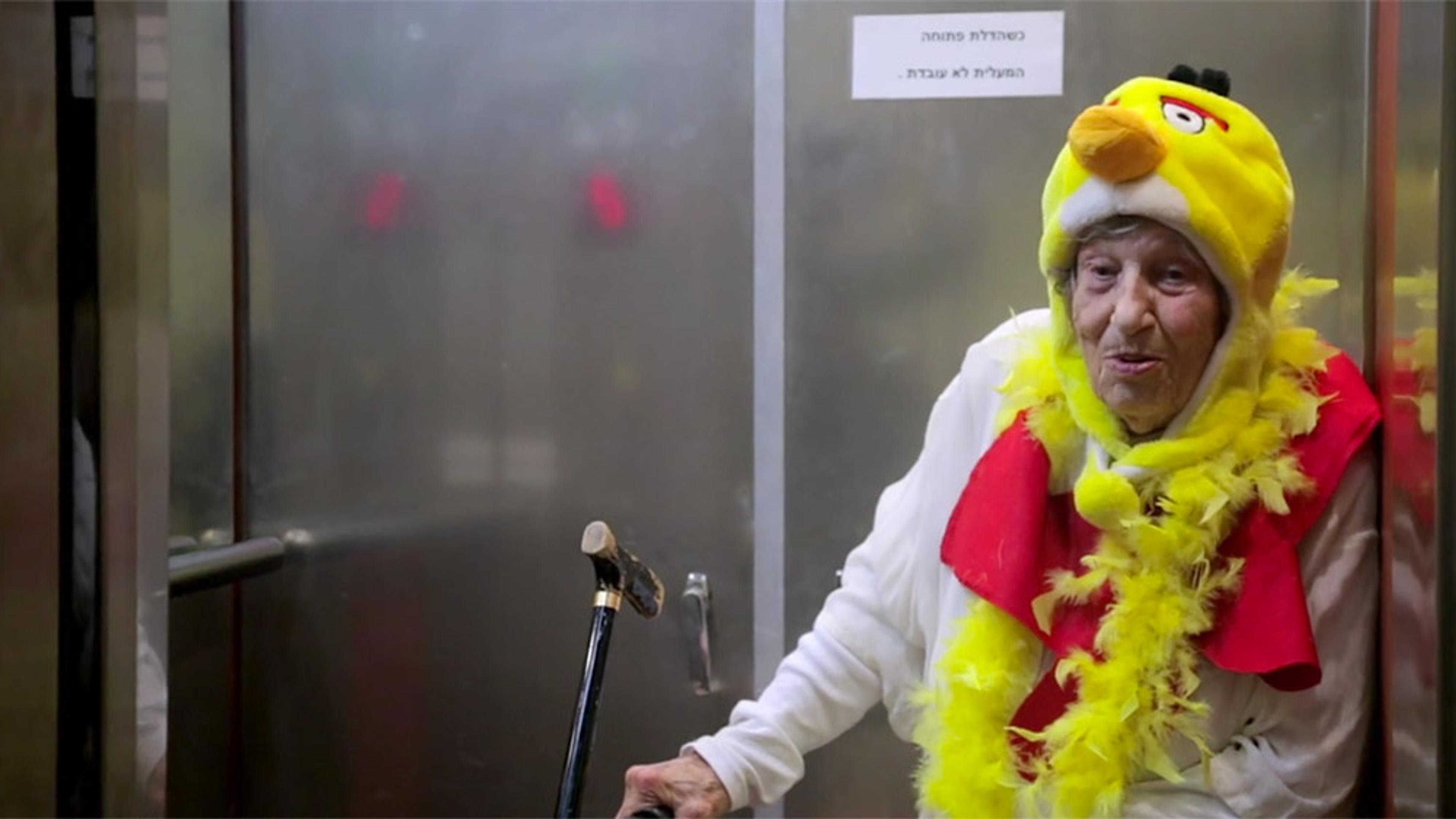 An elderly woman in a yellow bird costume with a walking stick, standing in front of a metal door with a sign in Hebrew.