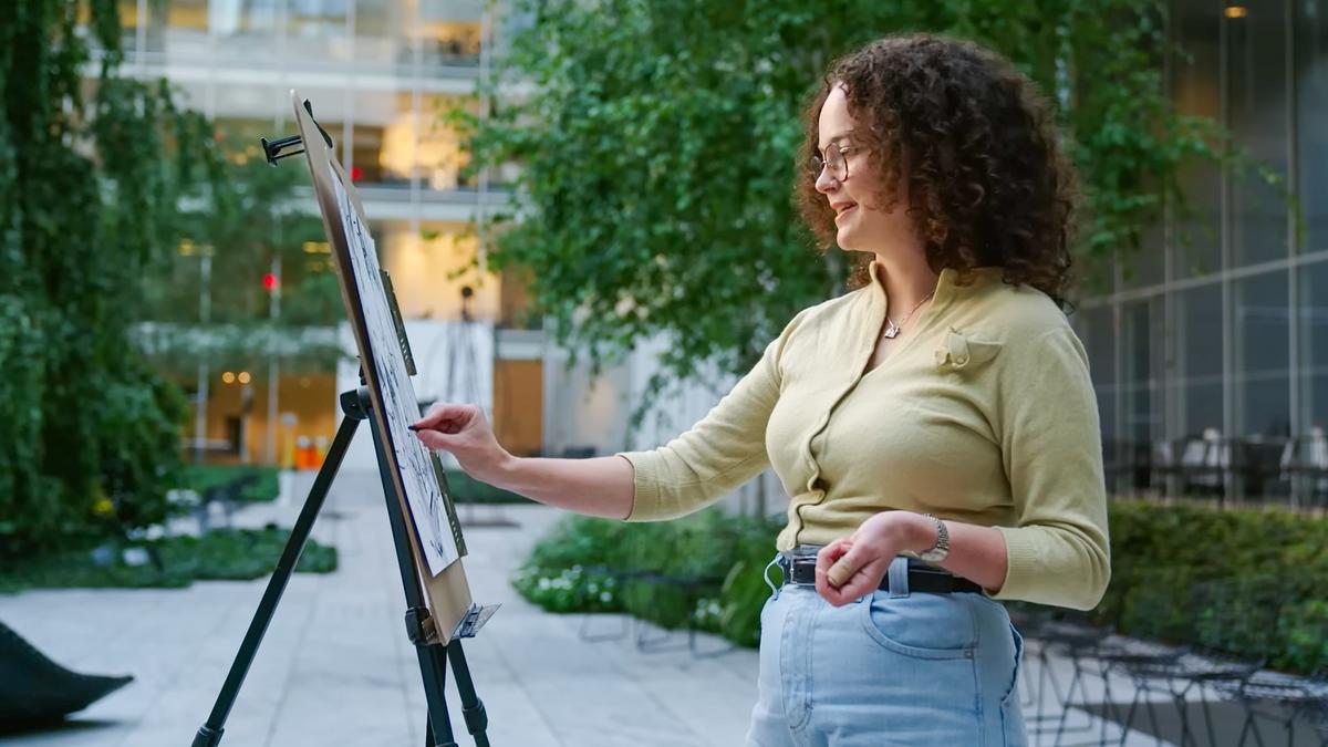 A woman drawing on an easel in a courtyard with greenery and a modern glass building in the background.