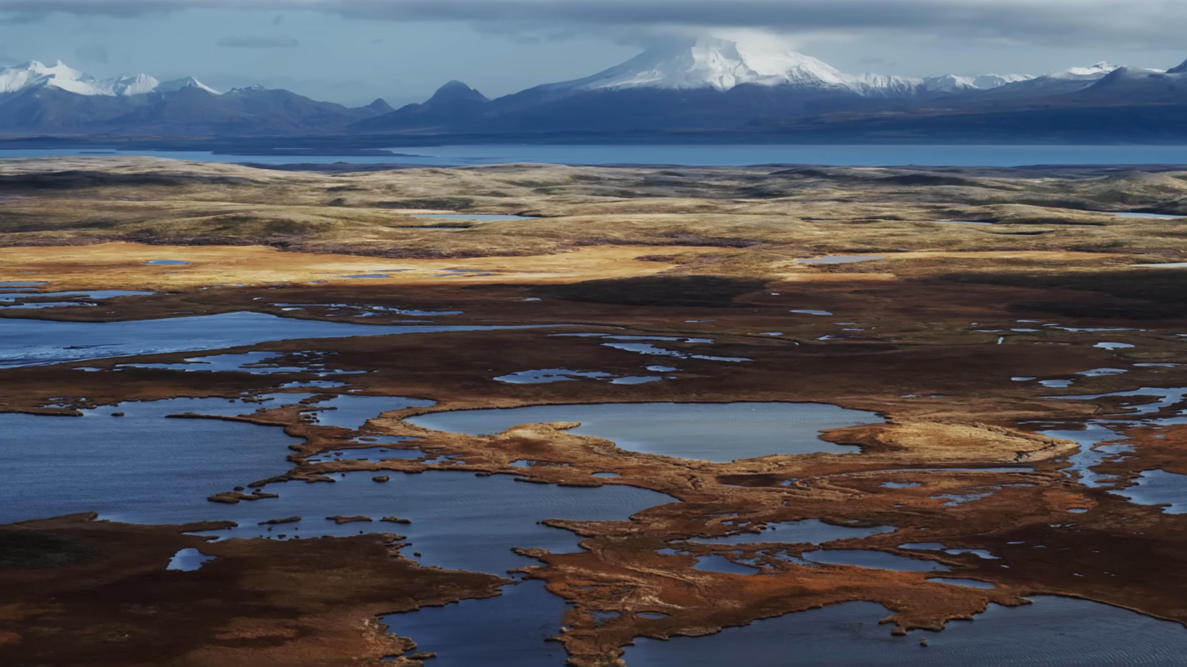 A landscape with lakes, tundra and distant snow-capped mountains under a cloudy sky.