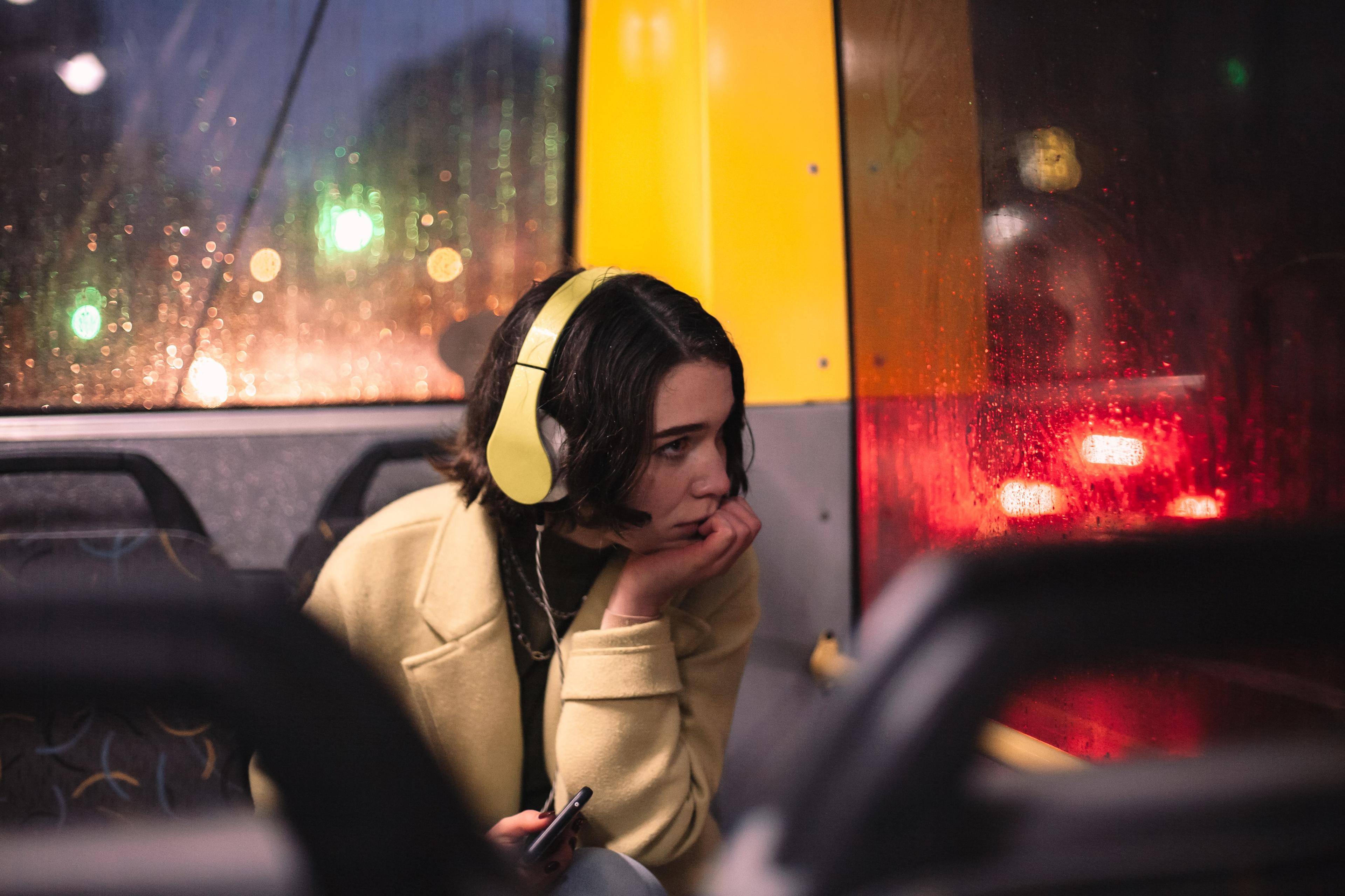 Photo of a woman with headphones on a rainy bus, looking thoughtful. Blurred city lights visible outside the window.