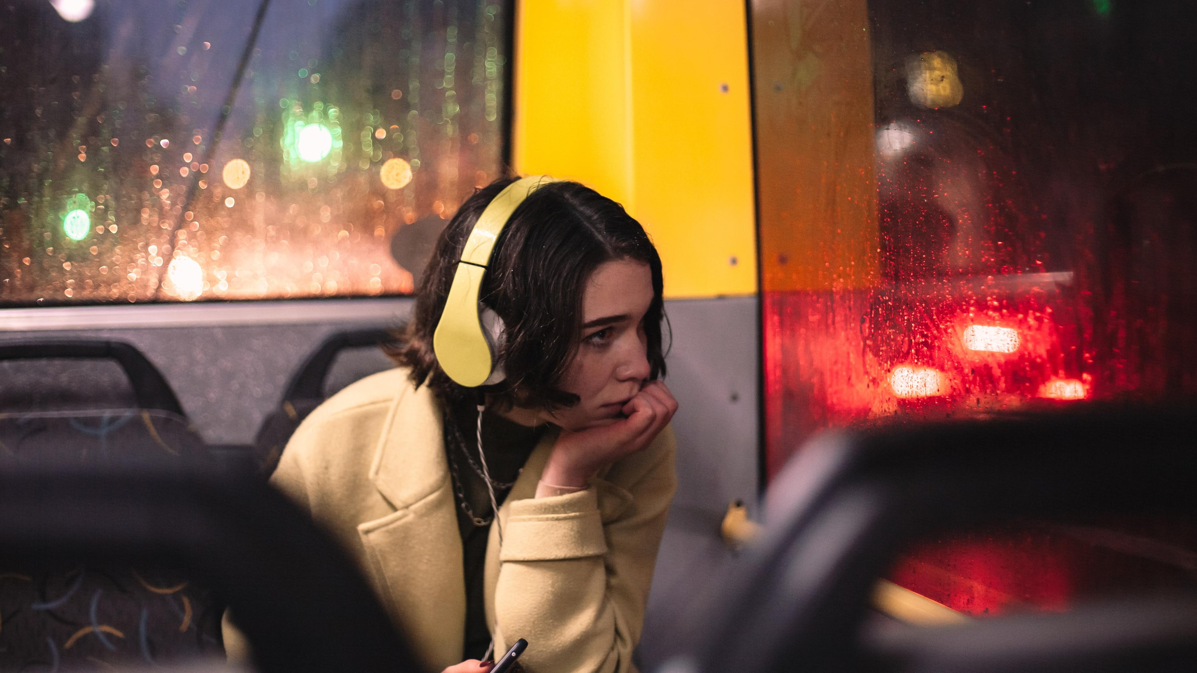 Photo of a woman with headphones on a rainy bus, looking thoughtful. Blurred city lights visible outside the window.