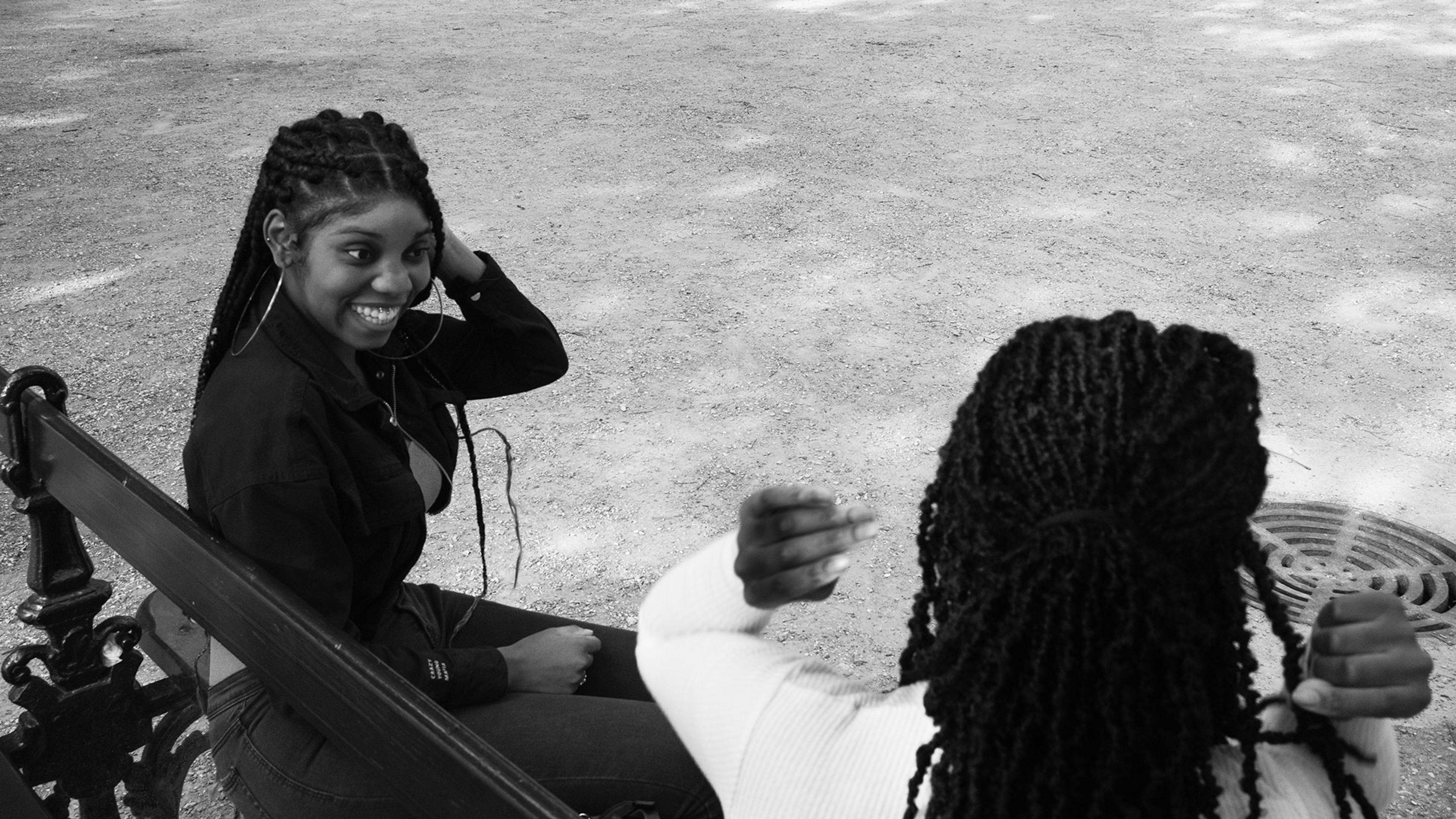 Two women with braided hair sitting on a park bench, smiling and engaging in conversation. One faces the camera, while the other is seen from behind.