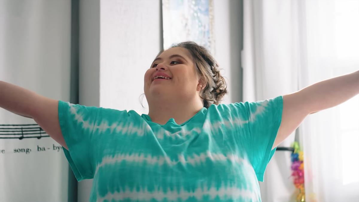 Photo of a person smiling with arms outstretched in a turquoise tie-dye shirt in a bright room.