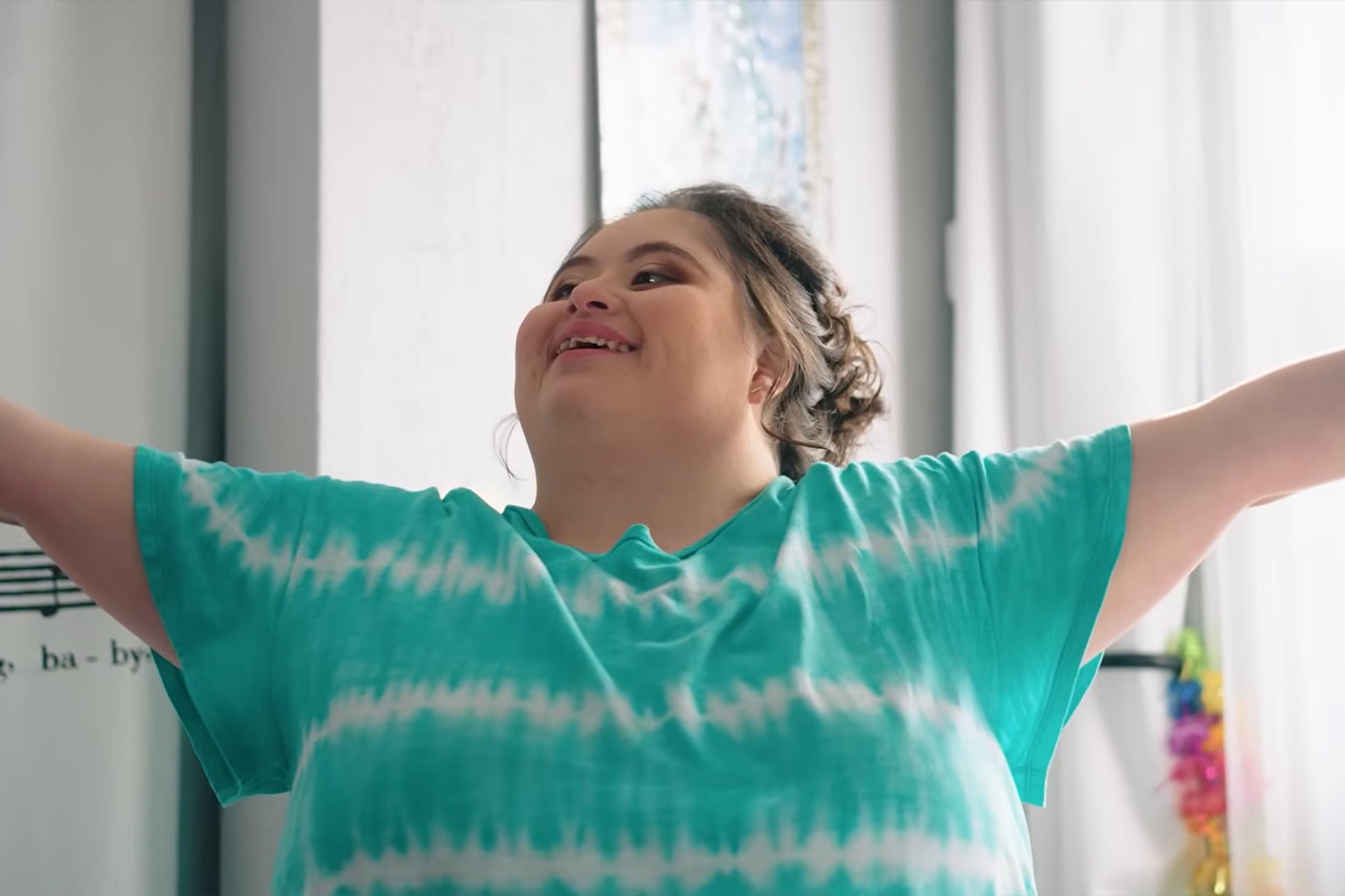 Photo of a person smiling with arms outstretched in a turquoise tie-dye shirt in a bright room.