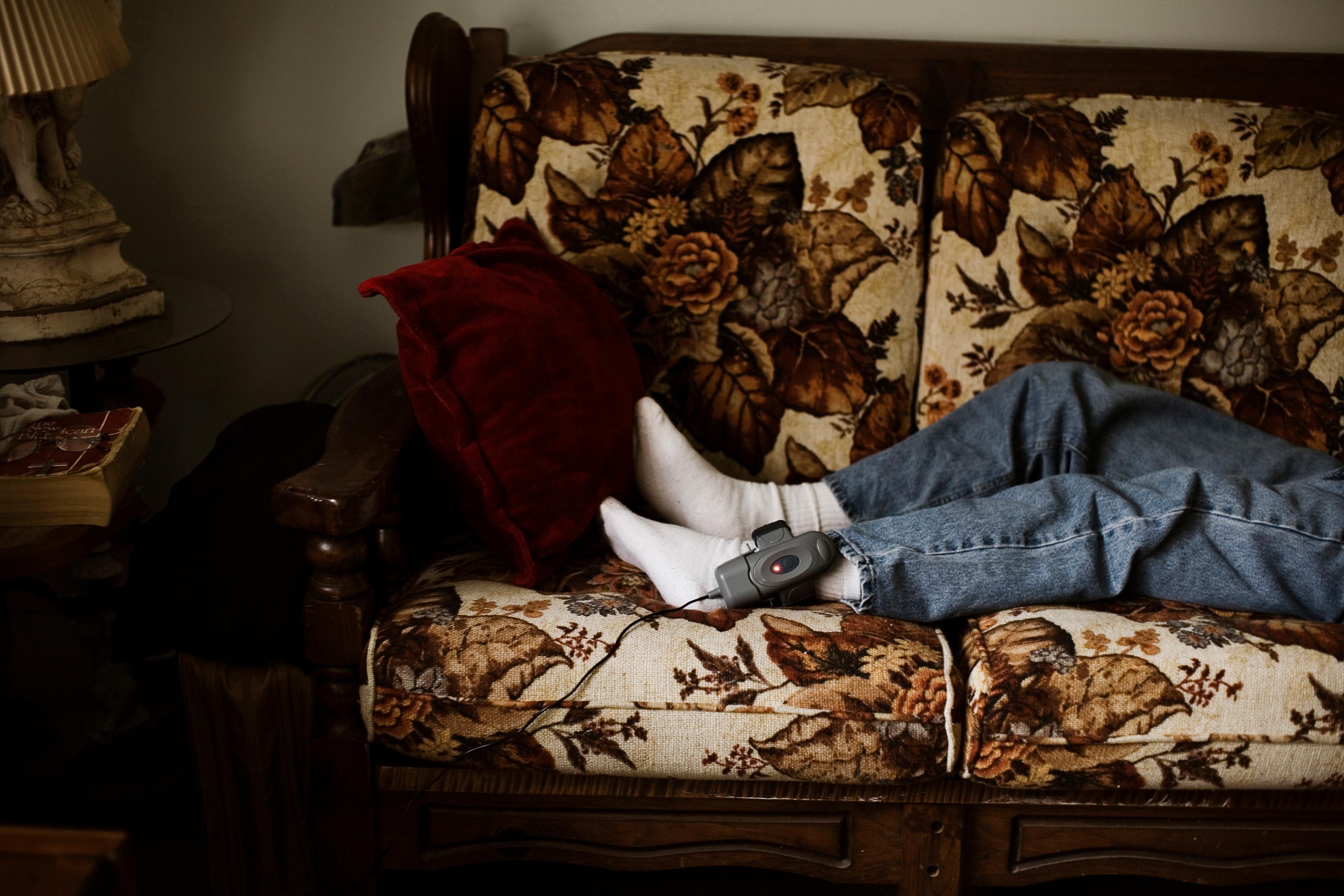 Photograph of a person lying on a floral sofa wearing jeans socks and an ankle monitor with a red cushion nearby.