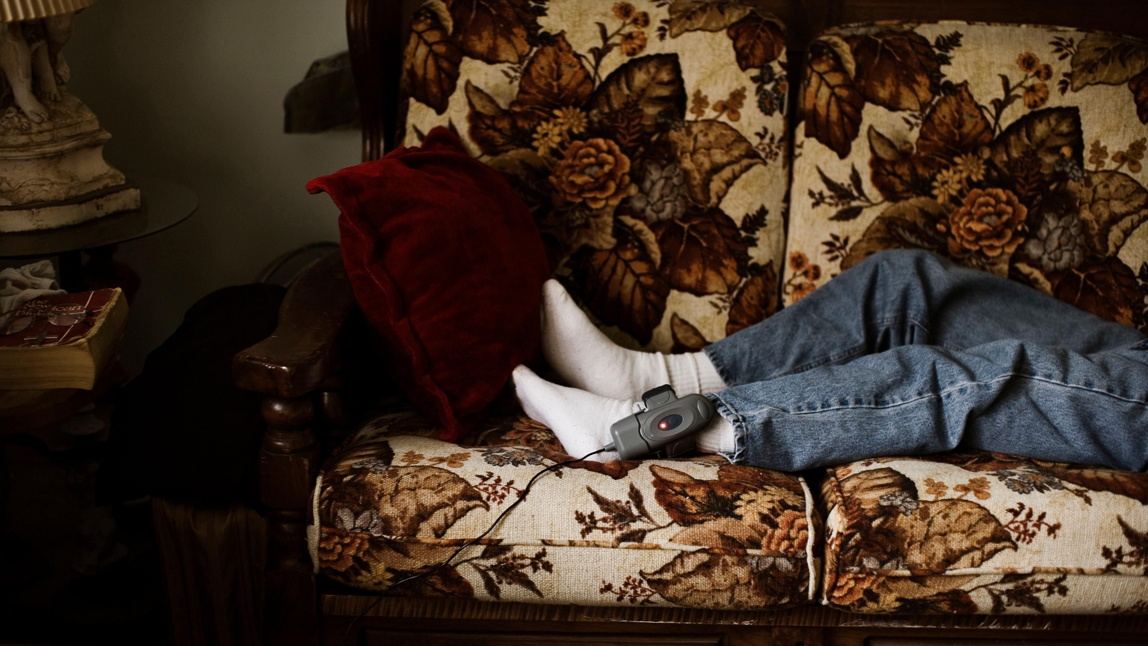 Photograph of a person lying on a floral sofa wearing jeans socks and an ankle monitor with a red cushion nearby.