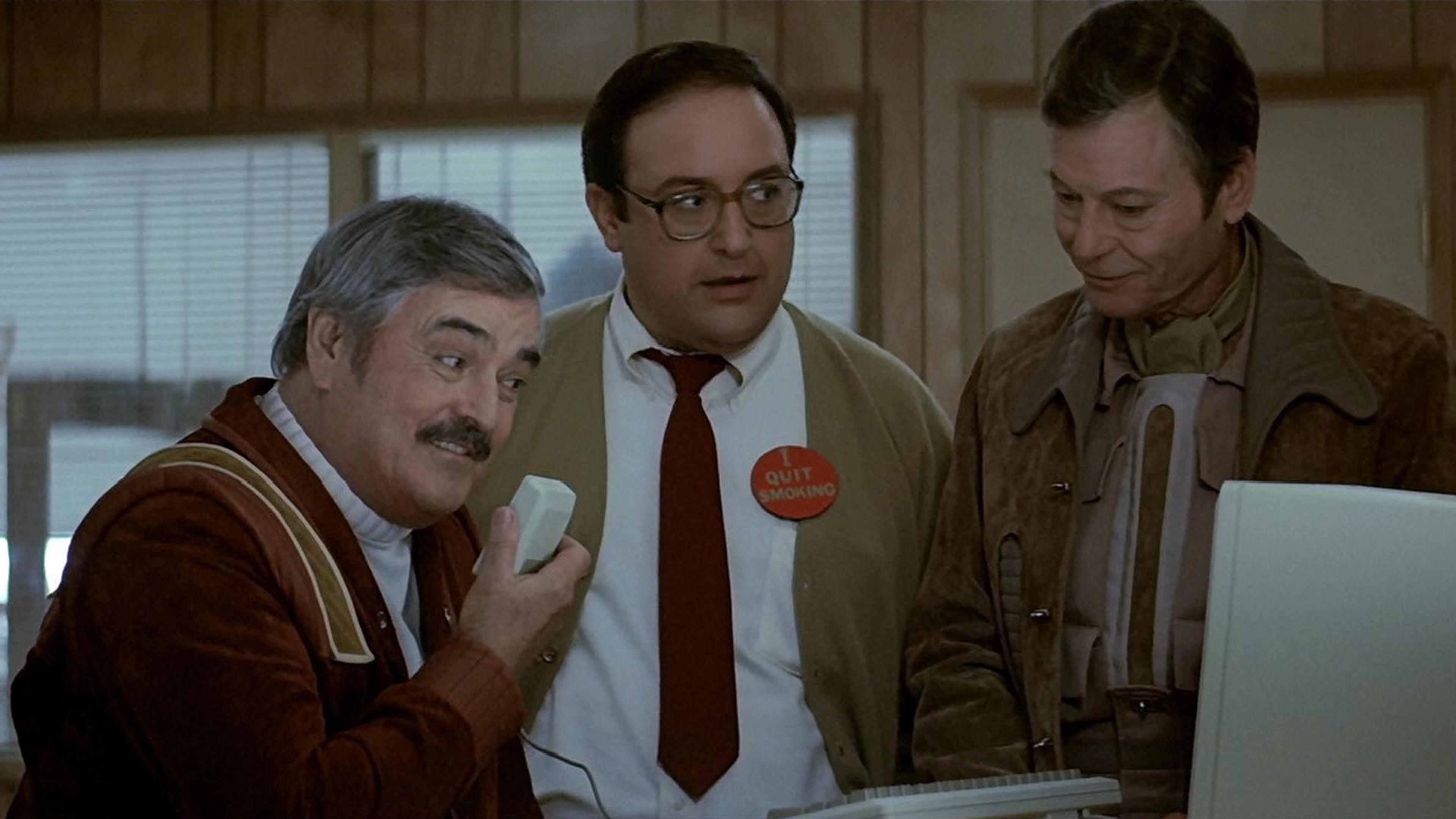 Scene from a film with three men indoors, one holding a computer mouse. A badge reads “I quit smoking.”