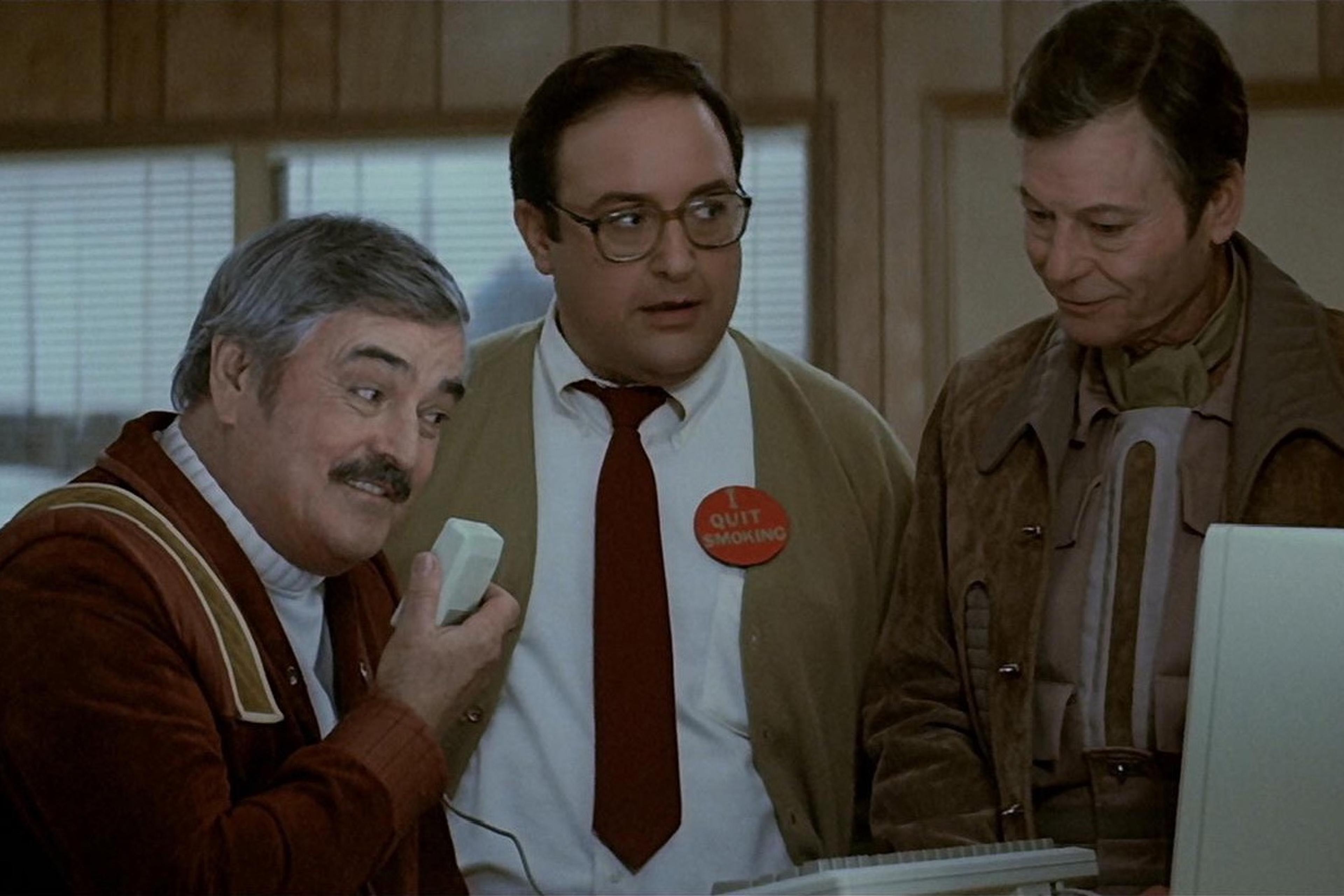 Scene from a film with three men indoors, one holding a computer mouse. A badge reads “I quit smoking.”