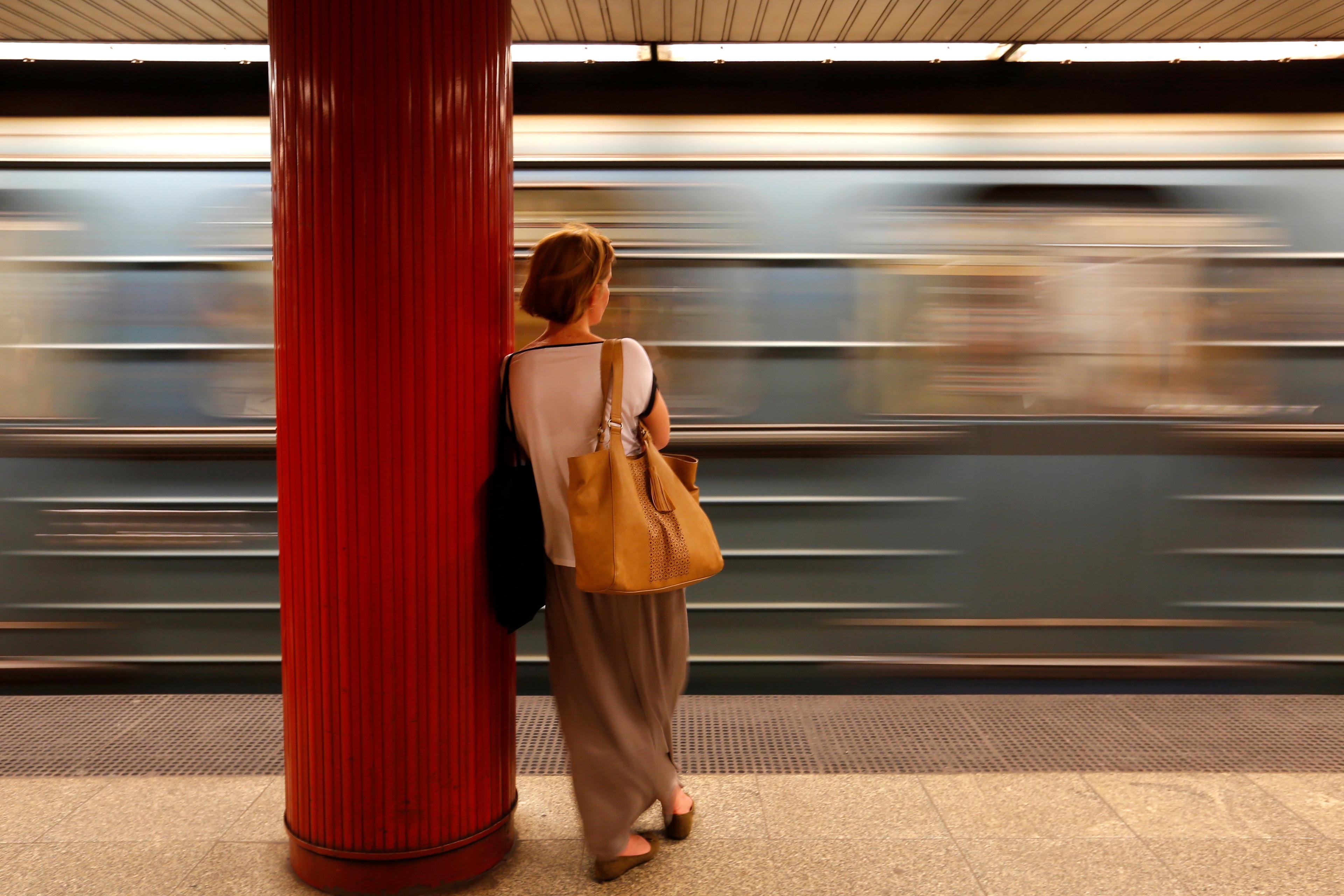 Photo of a woman leaning on a red pillar in a subway station as a train speeds by in a blur.
