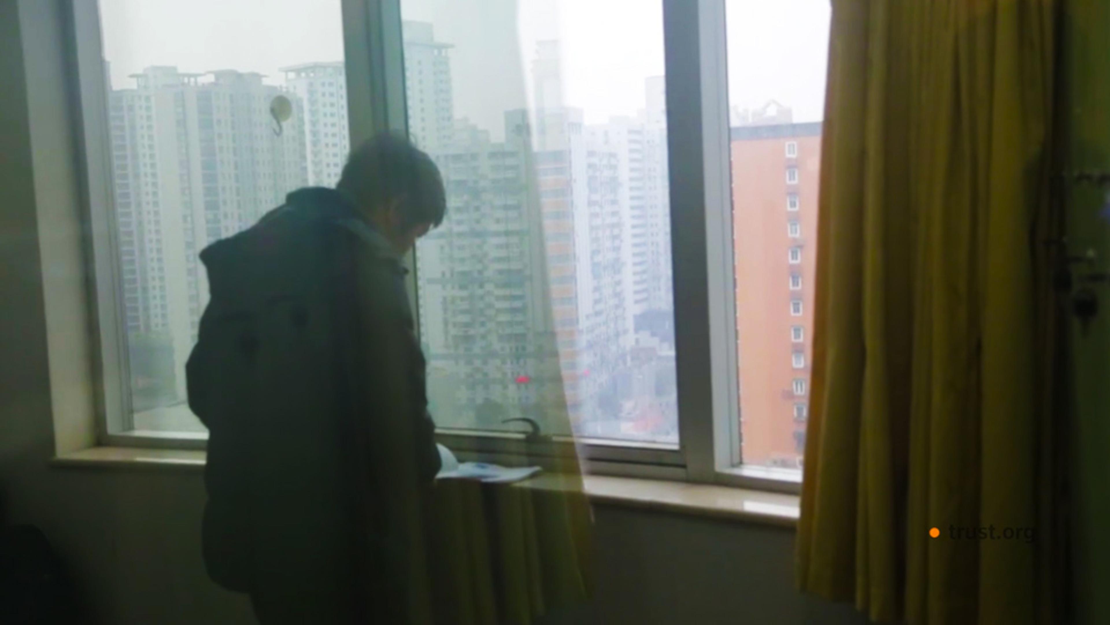 A person standing by a window in a high-rise apartment, looking at papers, with a cityscape visible outside.
