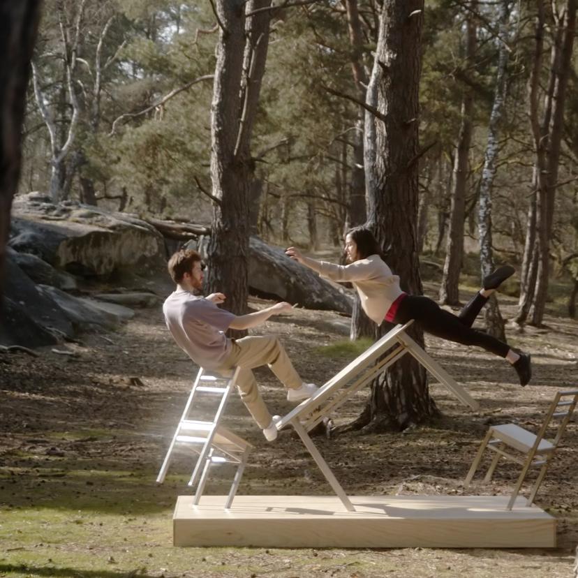 Two people in a park performing a balancing act on white chairs and a wooden plank, surrounded by pine trees.