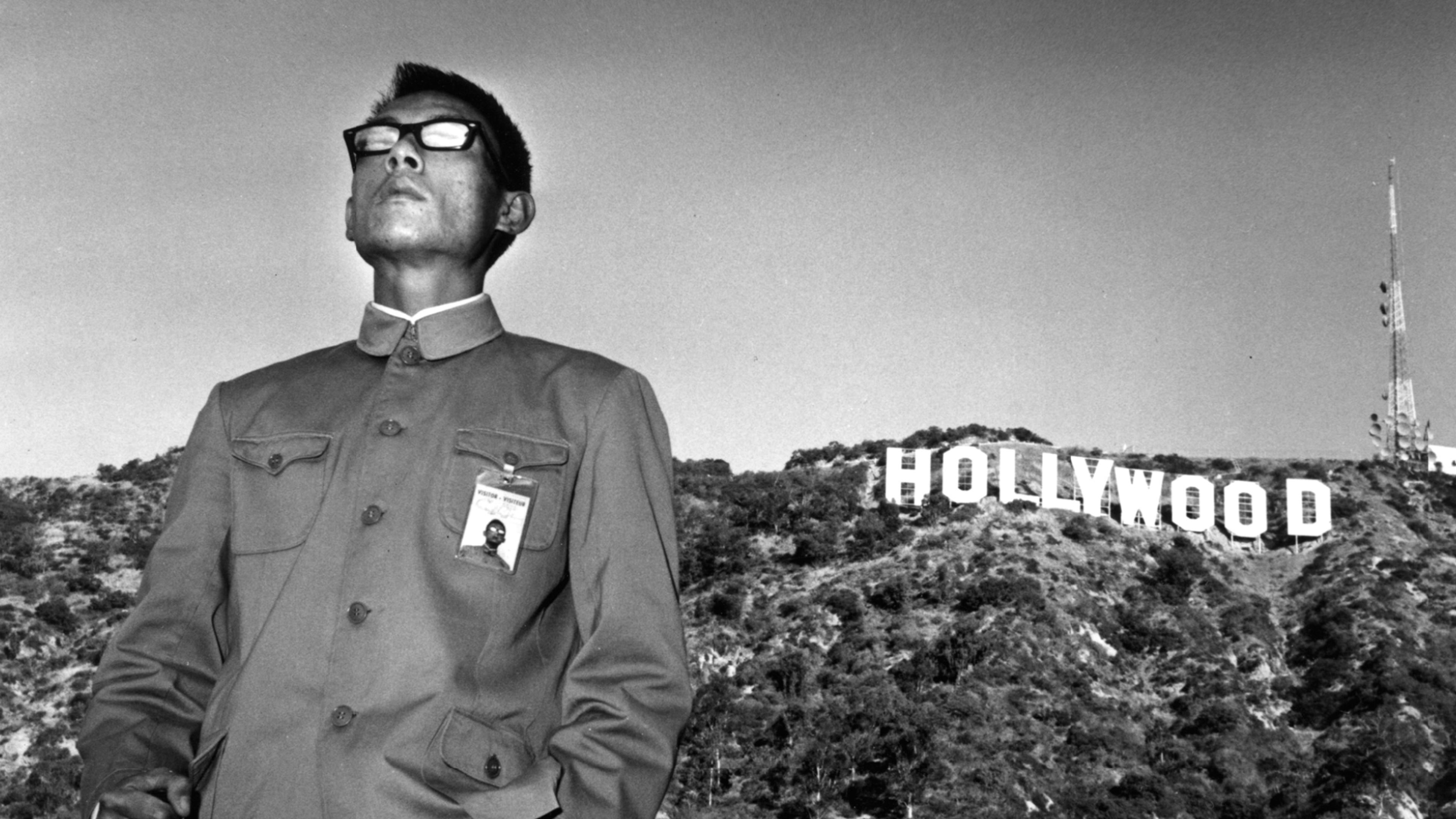 Black and white photo of a person in glasses and military attire standing in front of the Hollywood sign.