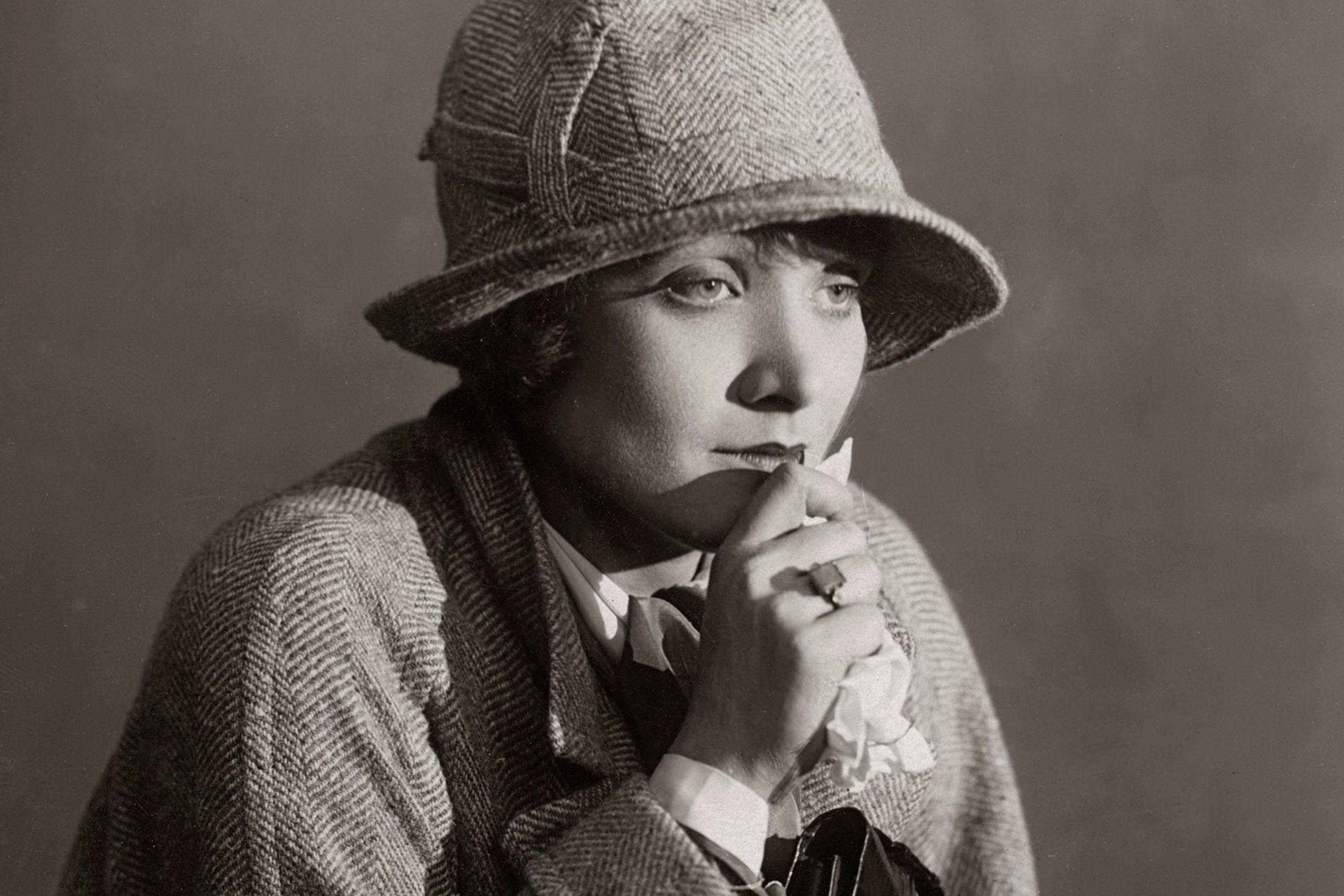 Black-and-white photo of a pensive woman in a herringbone coat and cloche hat, holding a handkerchief to her lips.
