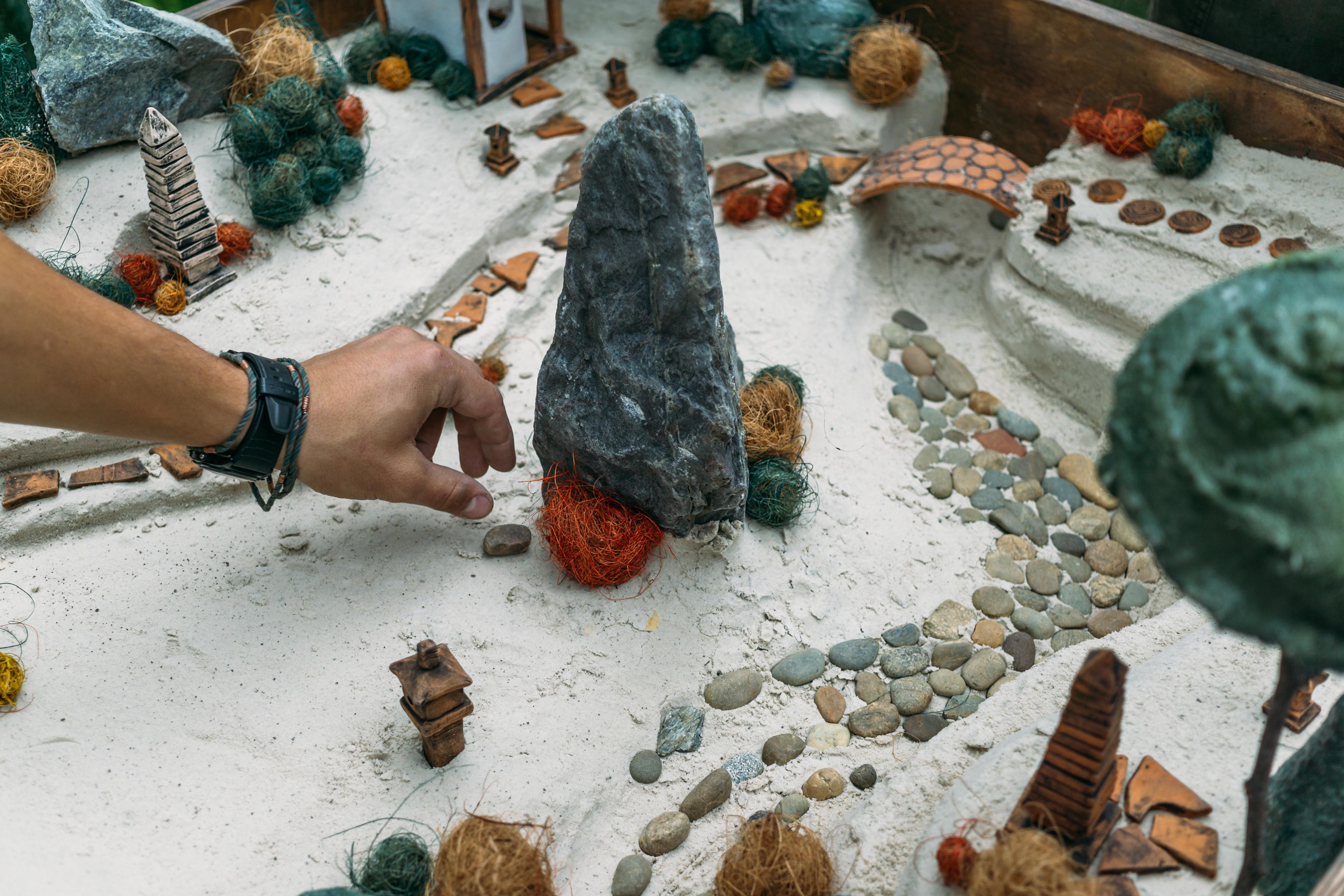 Miniature garden diorama with sand paths, small rocks, and a hand adjusting elements, captured in a photo.