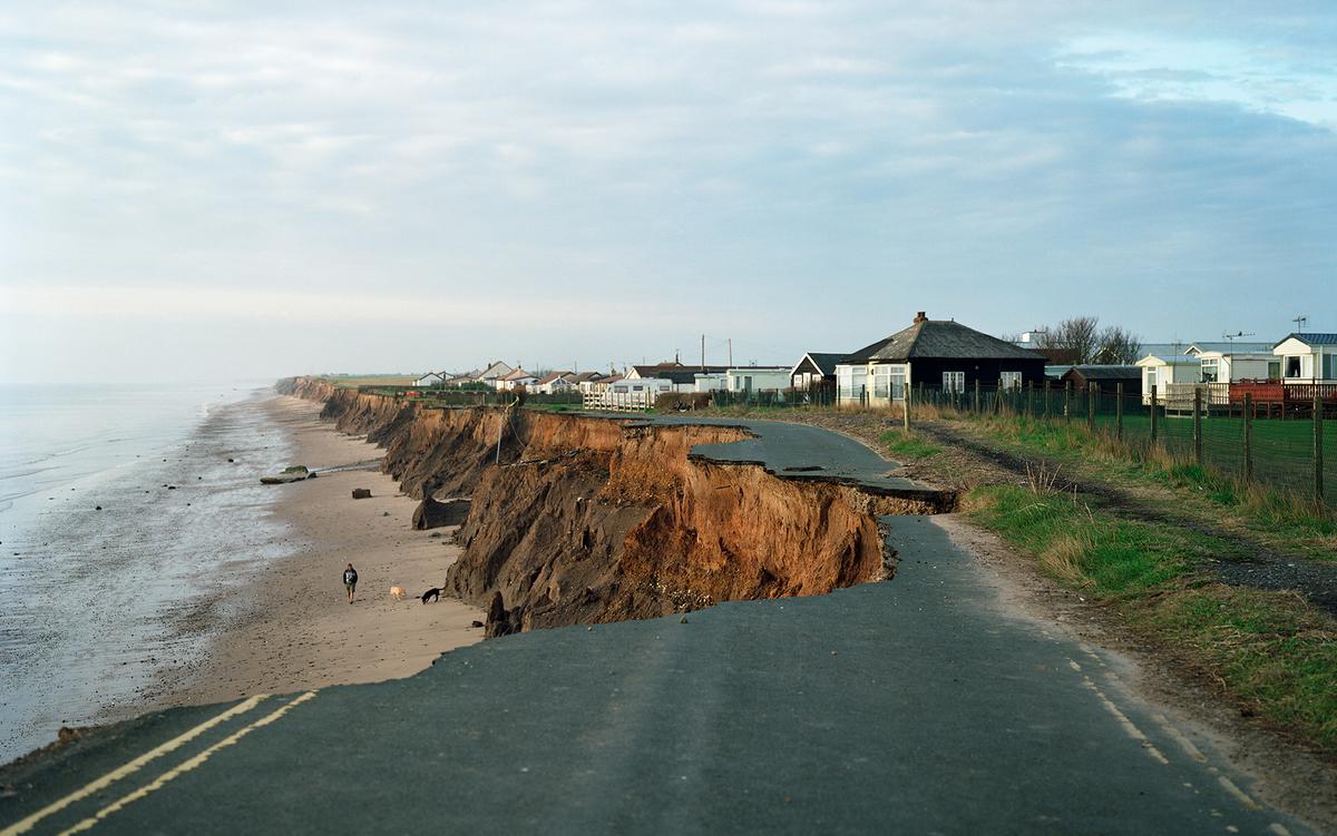 Coastal erosion showing a collapsed road beside the sea with houses nearby, a person walking a dog on the beach.