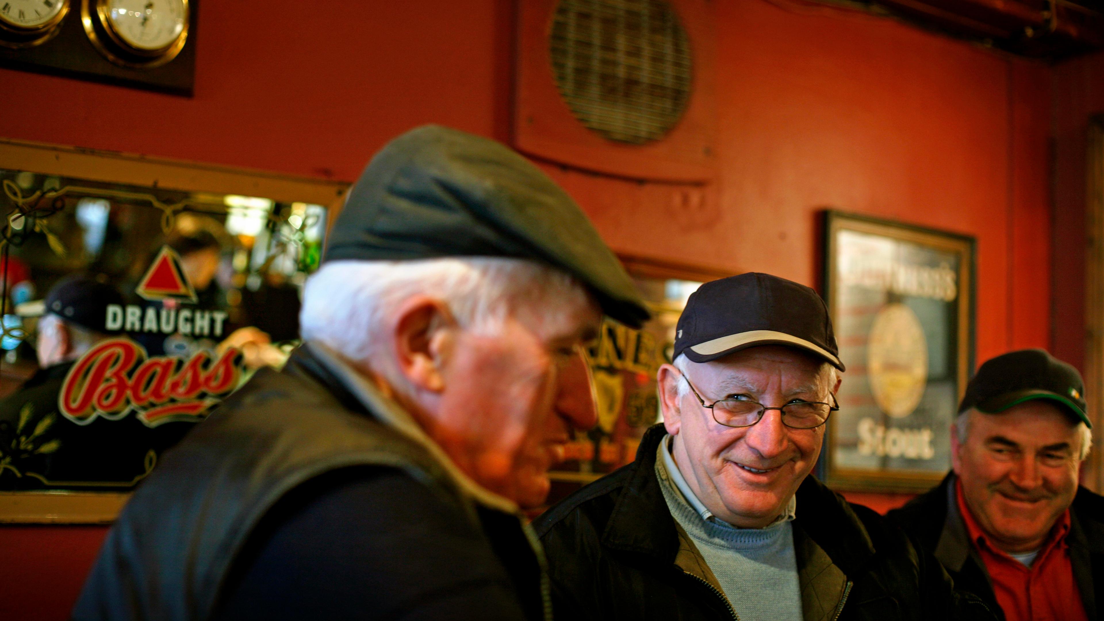 Photo of three older men in a pub, smiling and wearing caps. The background has pub mirrors and a red wall.
