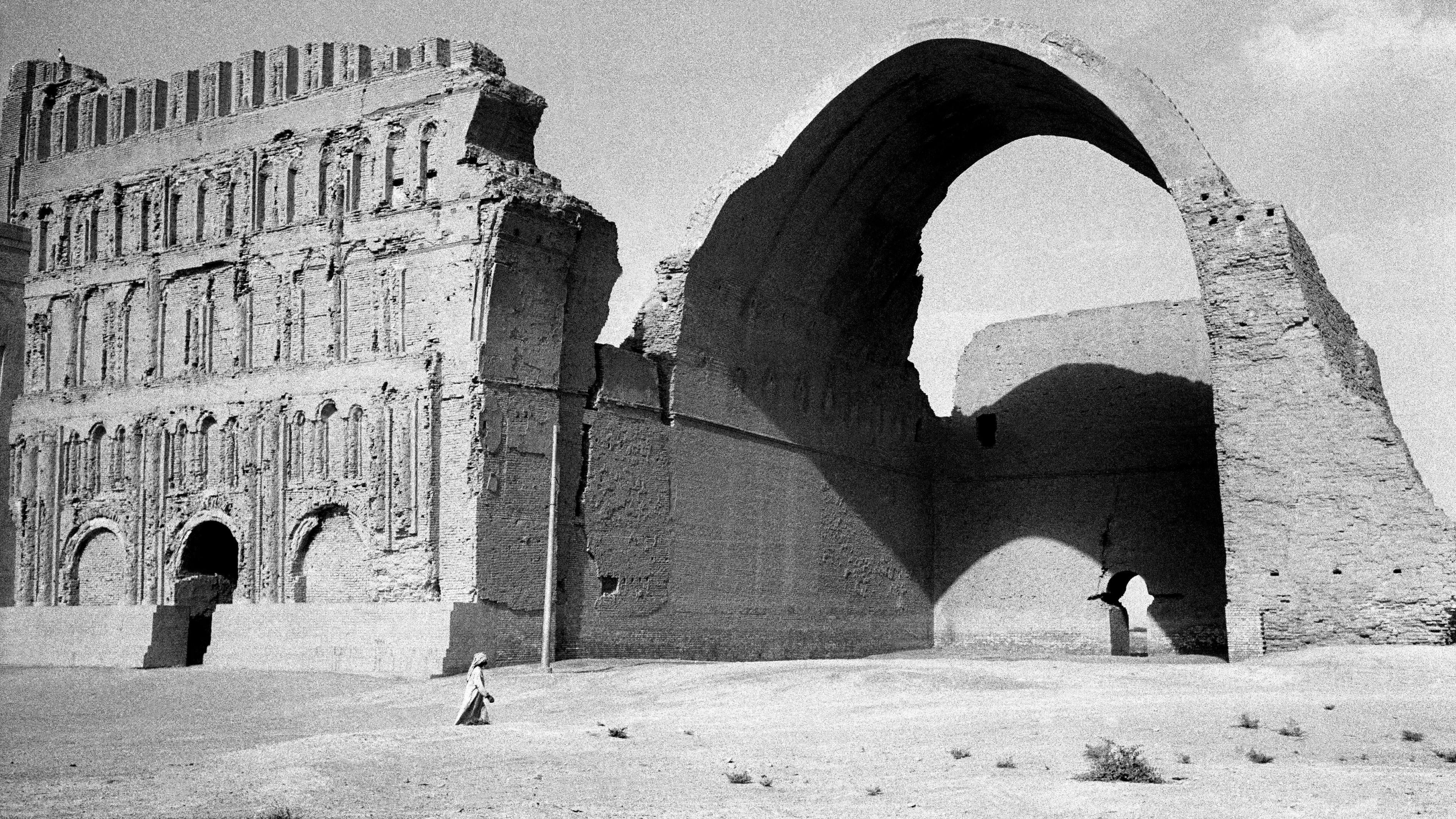 Black and white photo of ancient ruins with a large archway and detailed facade a lone figure walks in the foreground.