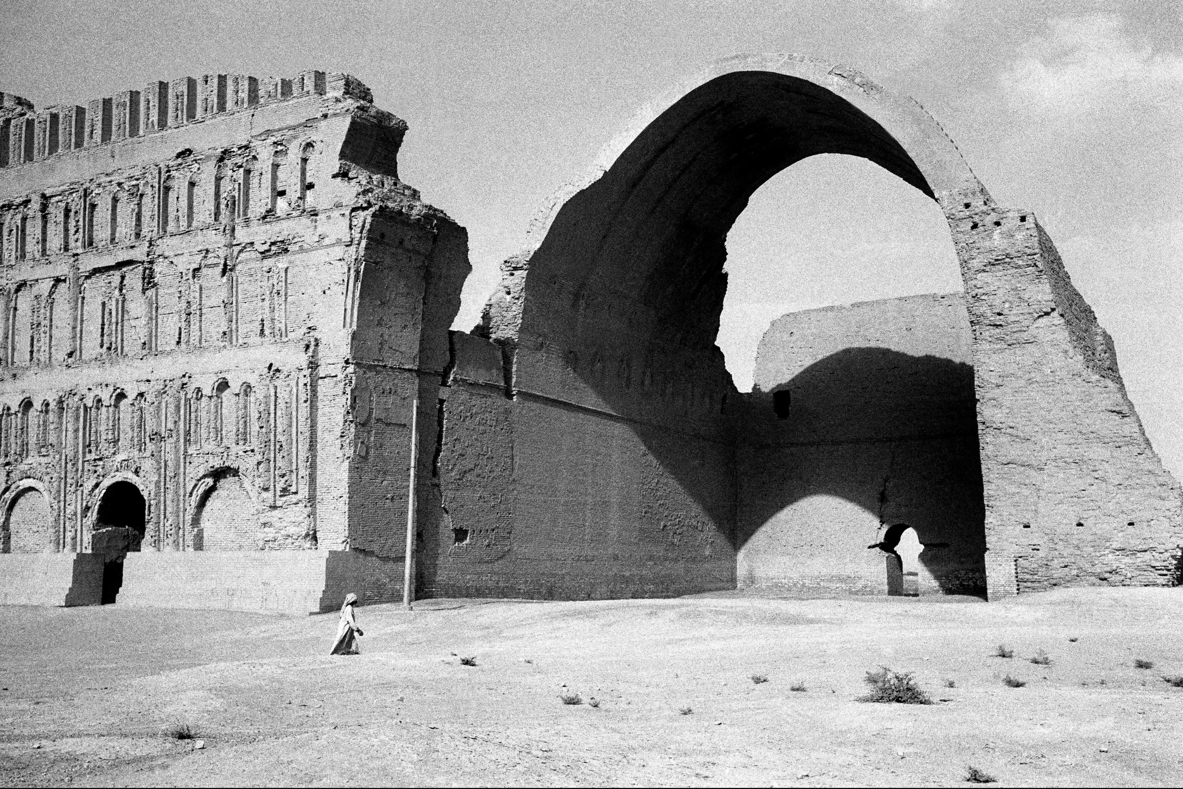 Black and white photo of ancient ruins with a large archway and detailed facade a lone figure walks in the foreground.