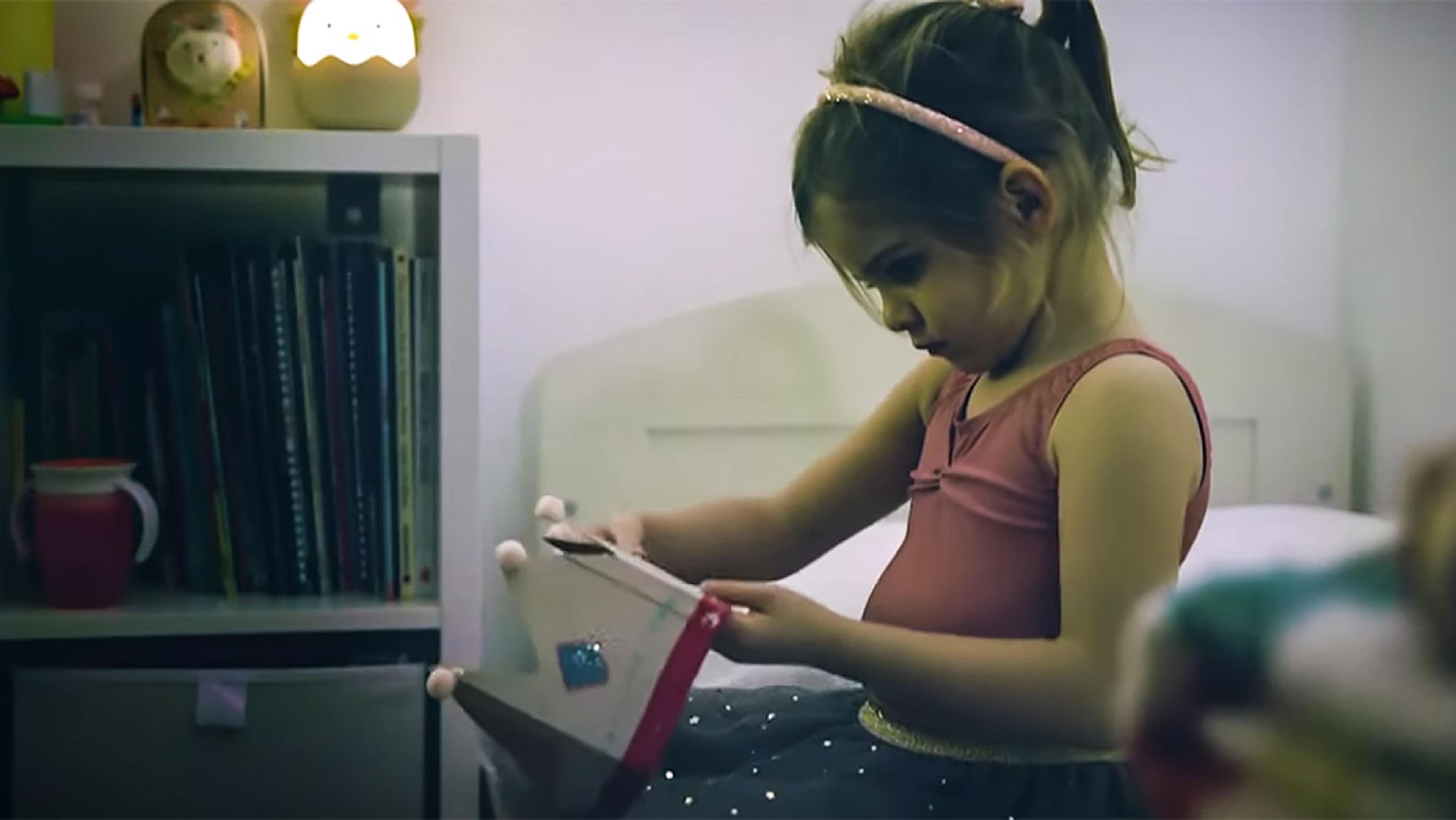 A young girl sitting on a bed wearing a pink dress with a headband playing with a paper crown in a bedroom with a bookshelf.