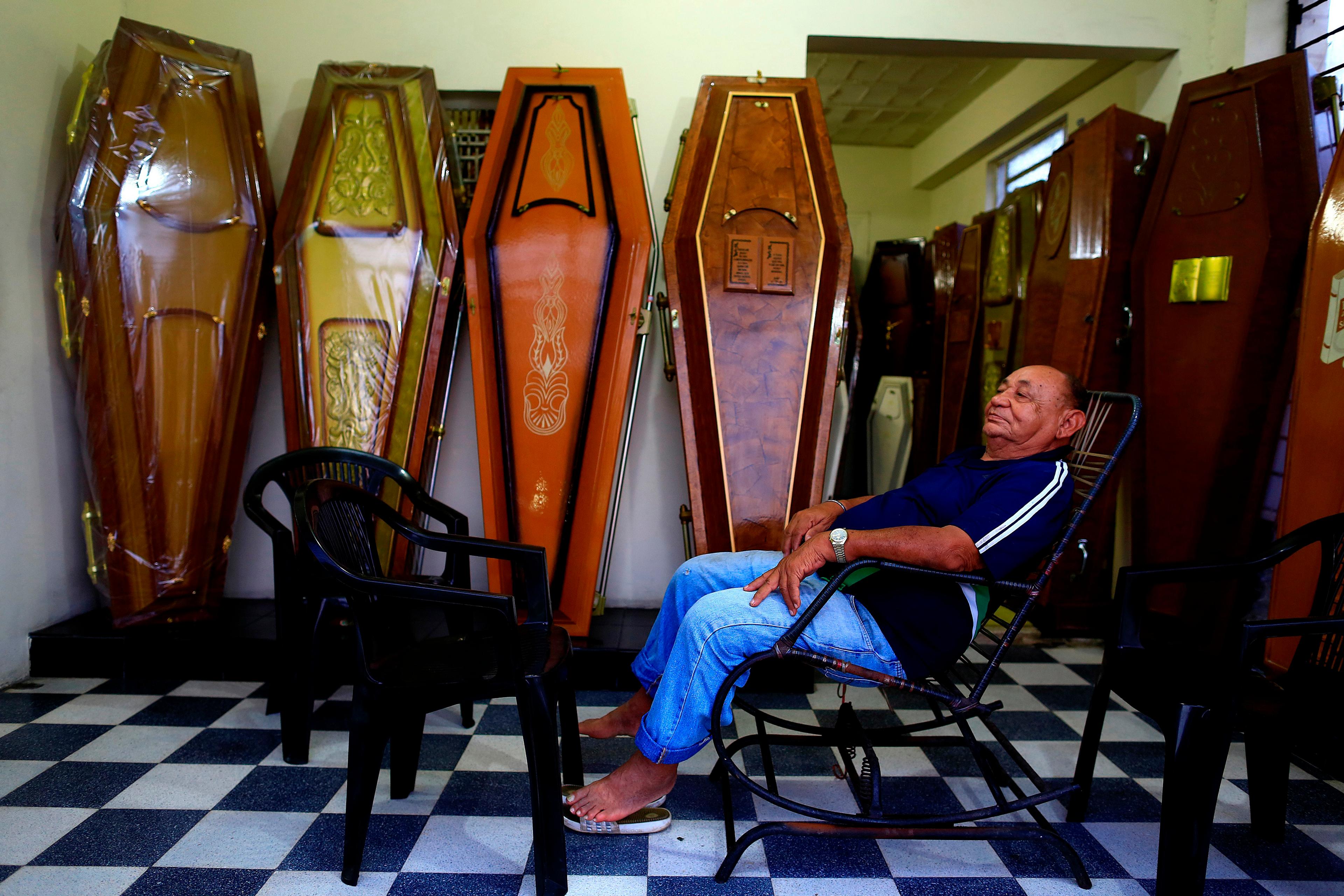 Photo of an elderly man sitting on a chair surrounded by various ornate coffins in a room with a chequered floor.