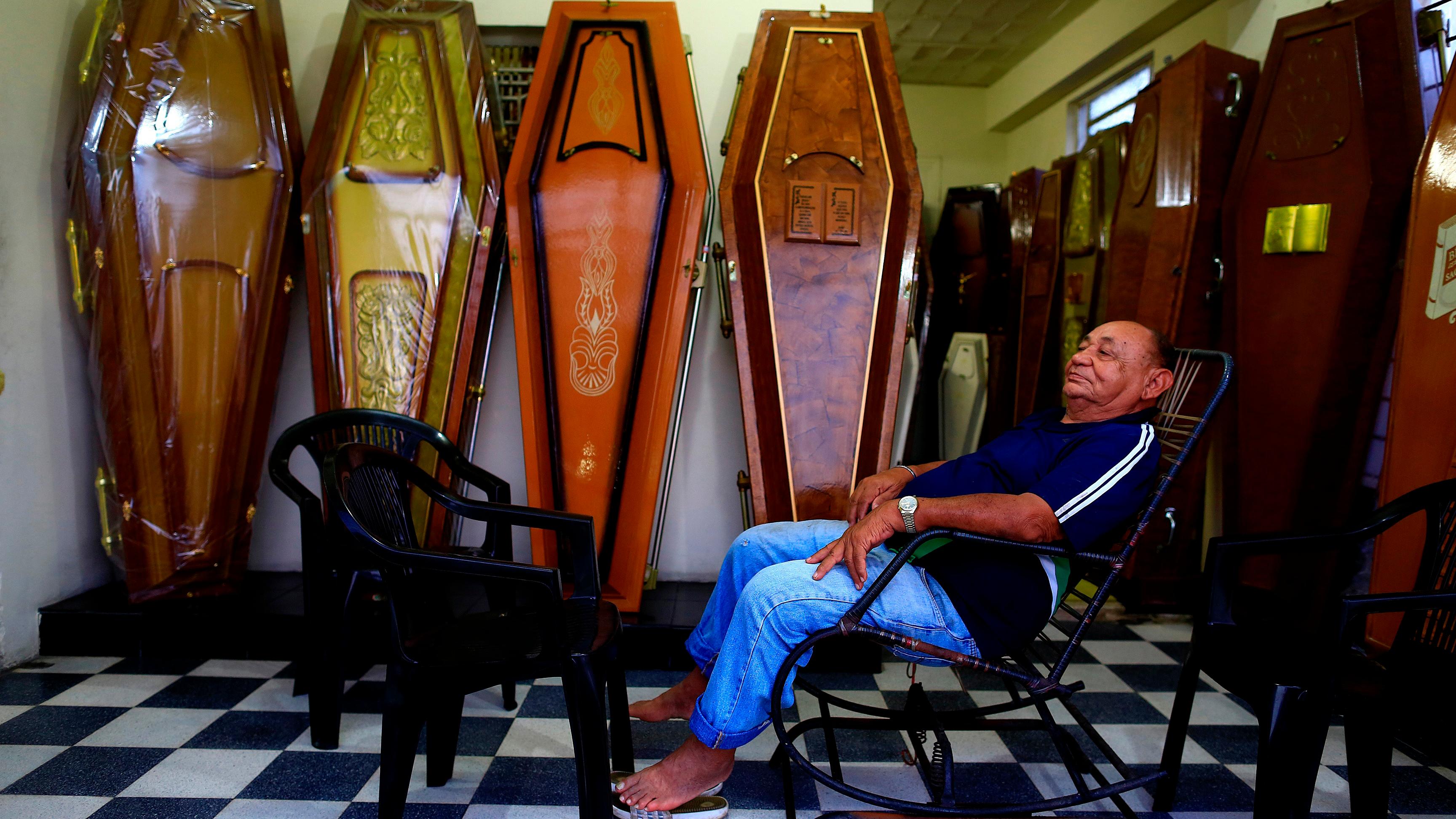 Photo of an elderly man sitting on a chair surrounded by various ornate coffins in a room with a chequered floor.