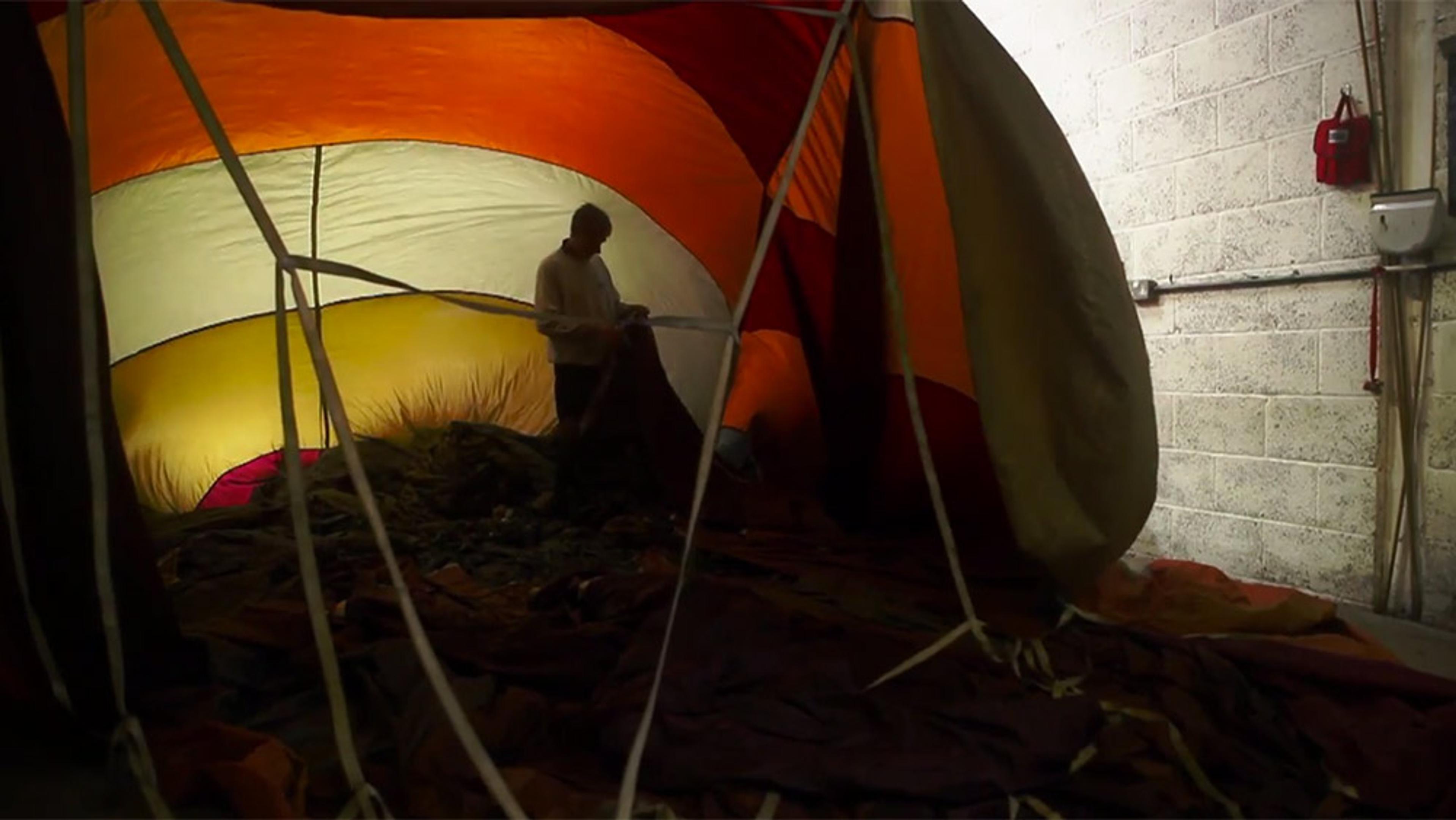 A person standing inside a colourful half-inflated hot air balloon near a white brick wall, illuminated from outside giving a warm glow.