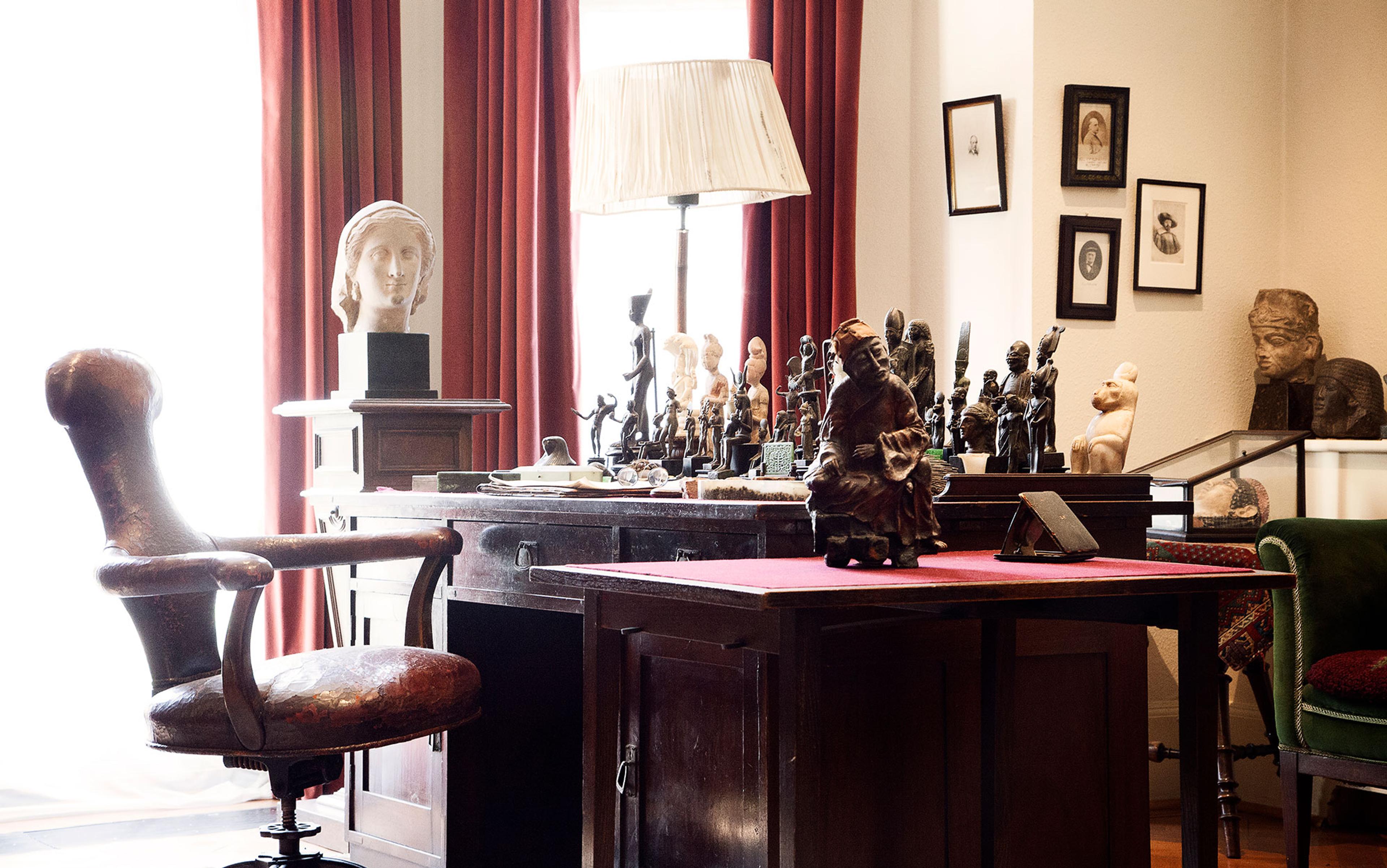 Photo of a study with a wooden desk, red curtains, bust sculptures and a leather chair in a well-lit room.