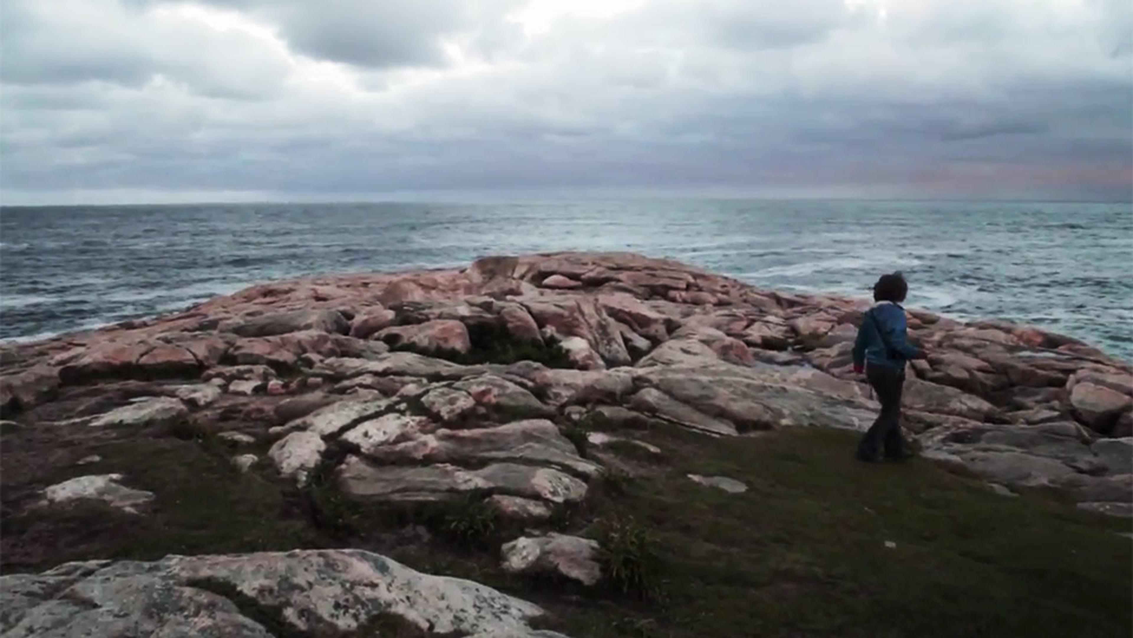 A person walking on rocky terrain by the sea under a cloudy sky. The horizon separates the turbulent water from the sky.