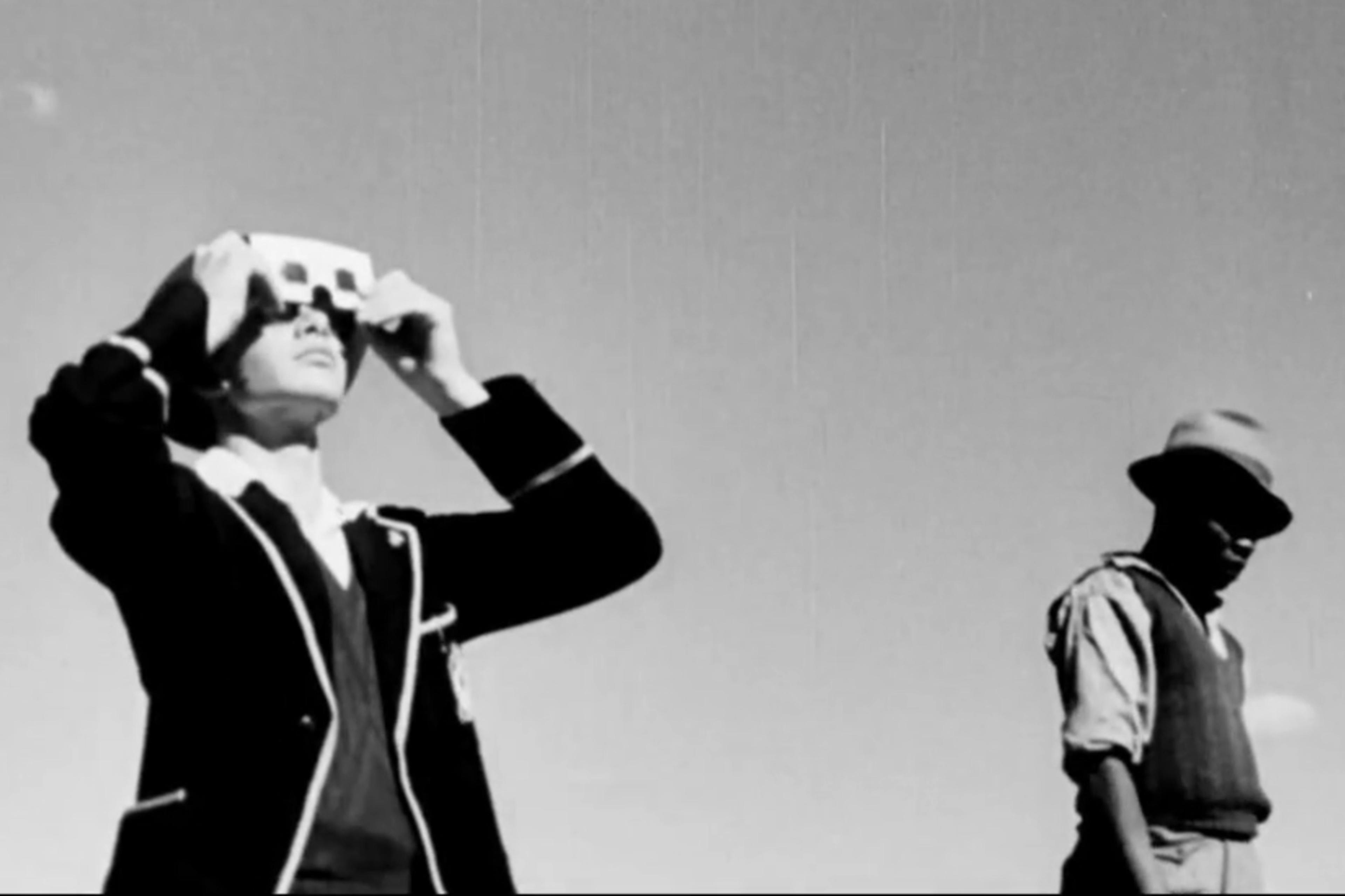 Black-and-white photo of two people; one holding binoculars to their eyes, the other wearing a hat and looking down.
