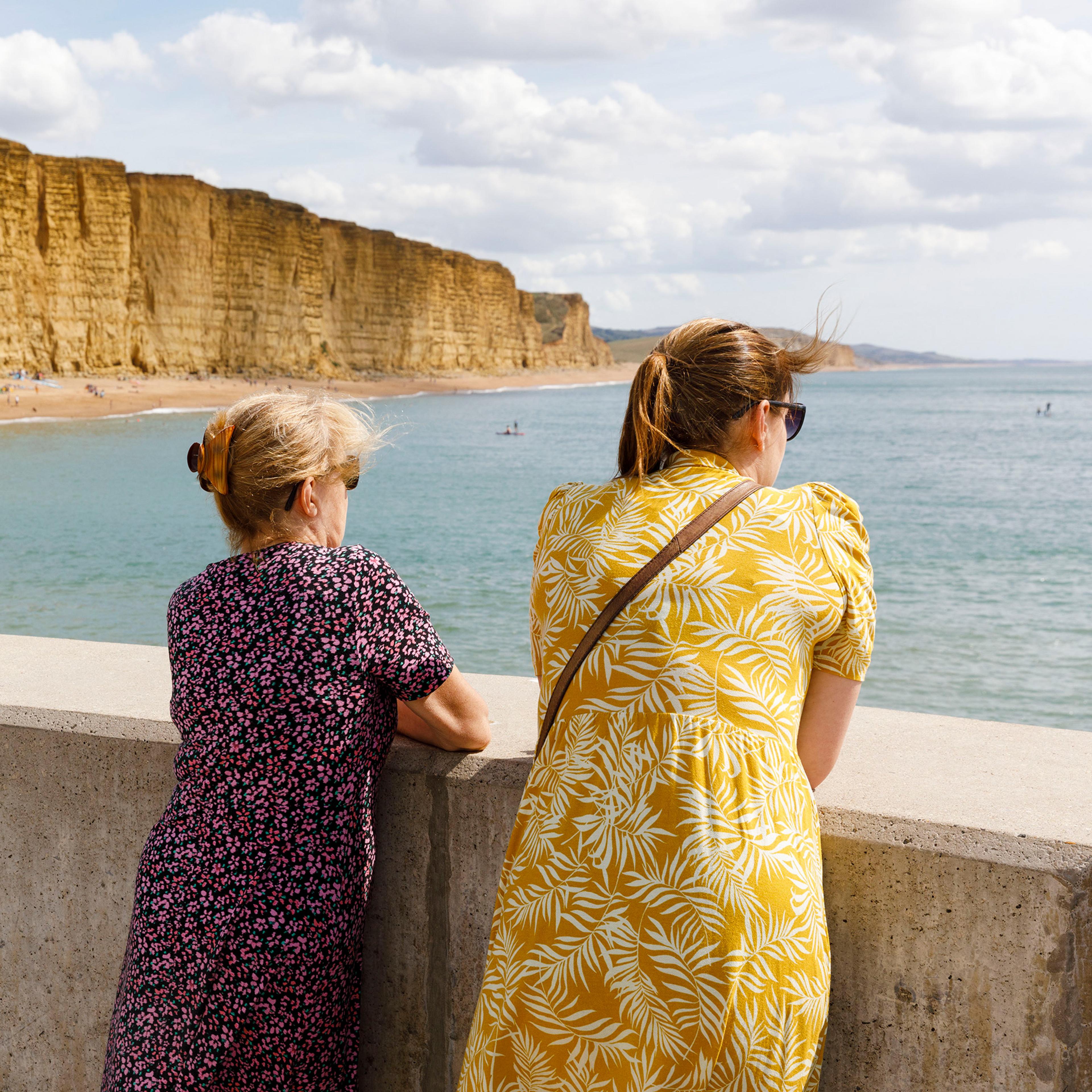 Two women in printed dresses stand at a concrete railing, overlooking a beach and cliffs with the sea stretching into the horizon.