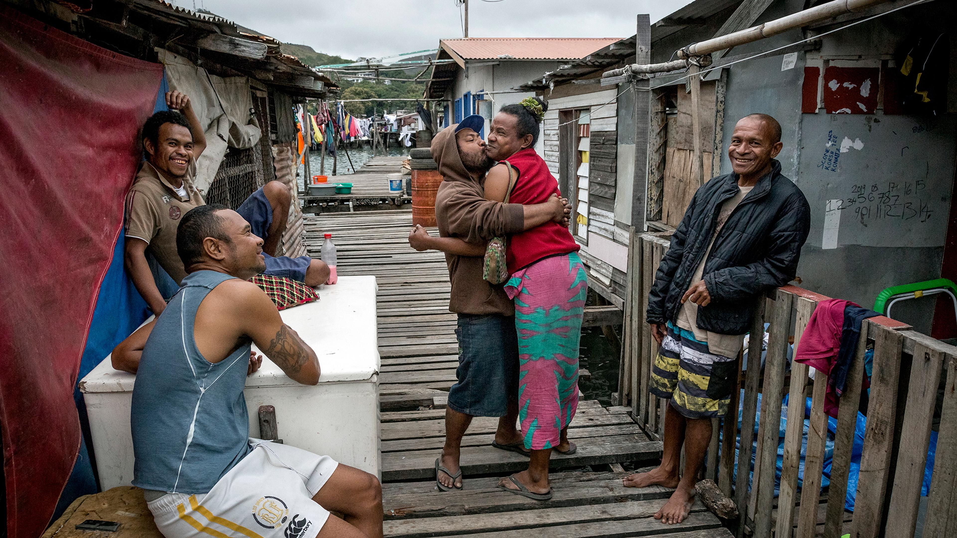 Photo of five people on a wooden walkway in a waterside village smiling, one pair embracing, colourful laundry in background.