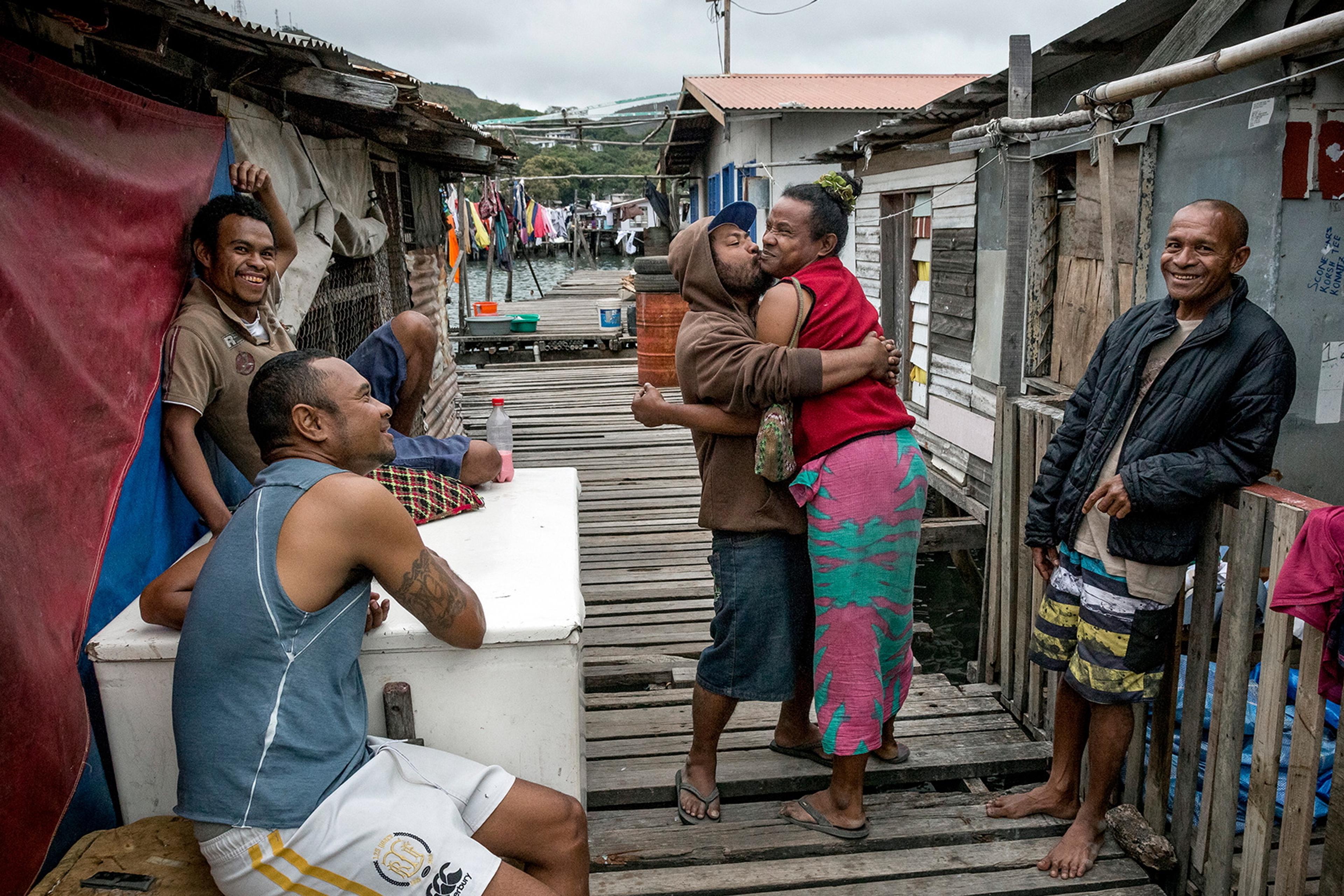 Photo of five people on a wooden walkway in a waterside village smiling, one pair embracing, colourful laundry in background.