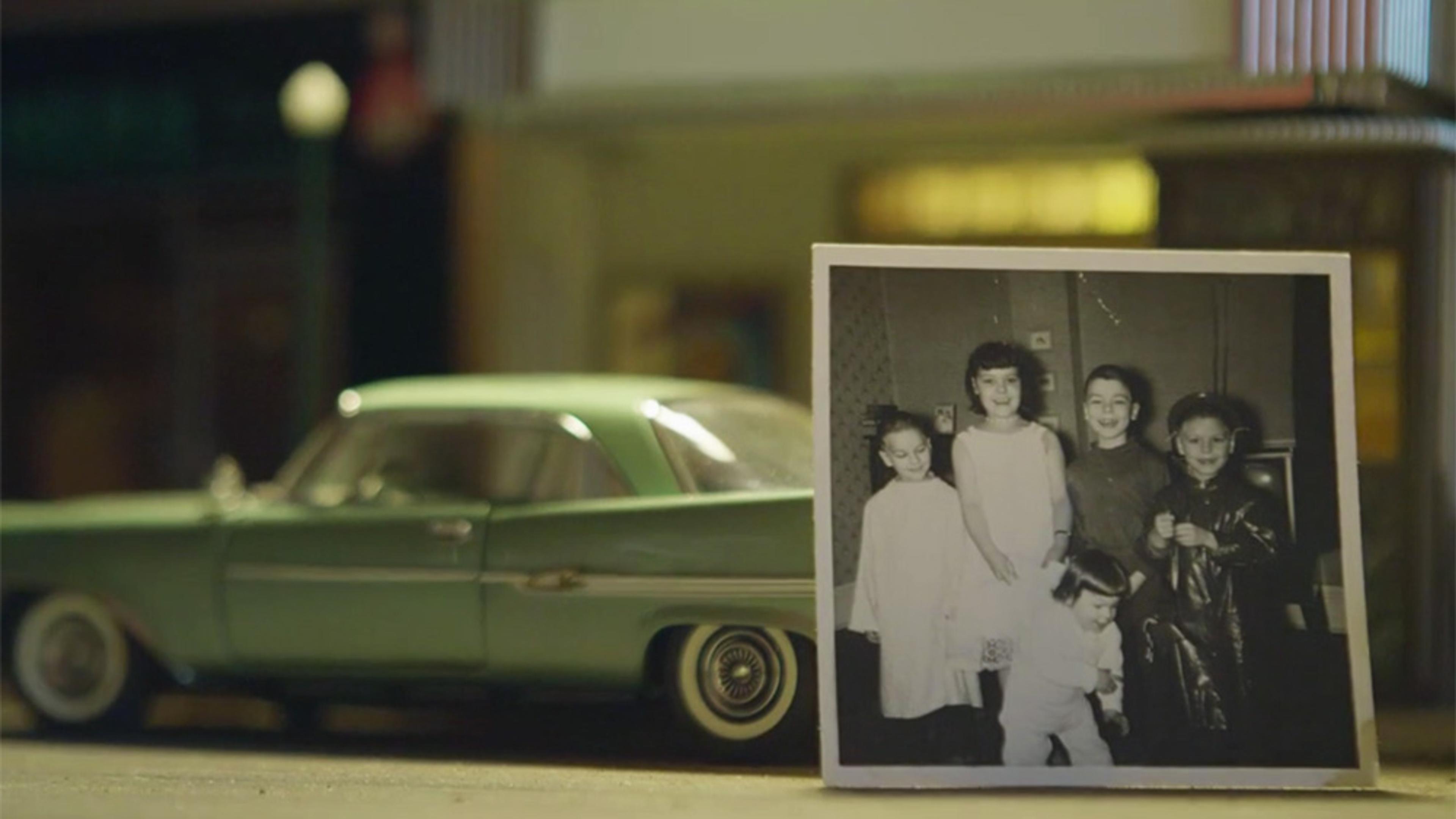 Photo of an old black-and-white picture showing five children, placed in front of a vintage green car on a city street background.