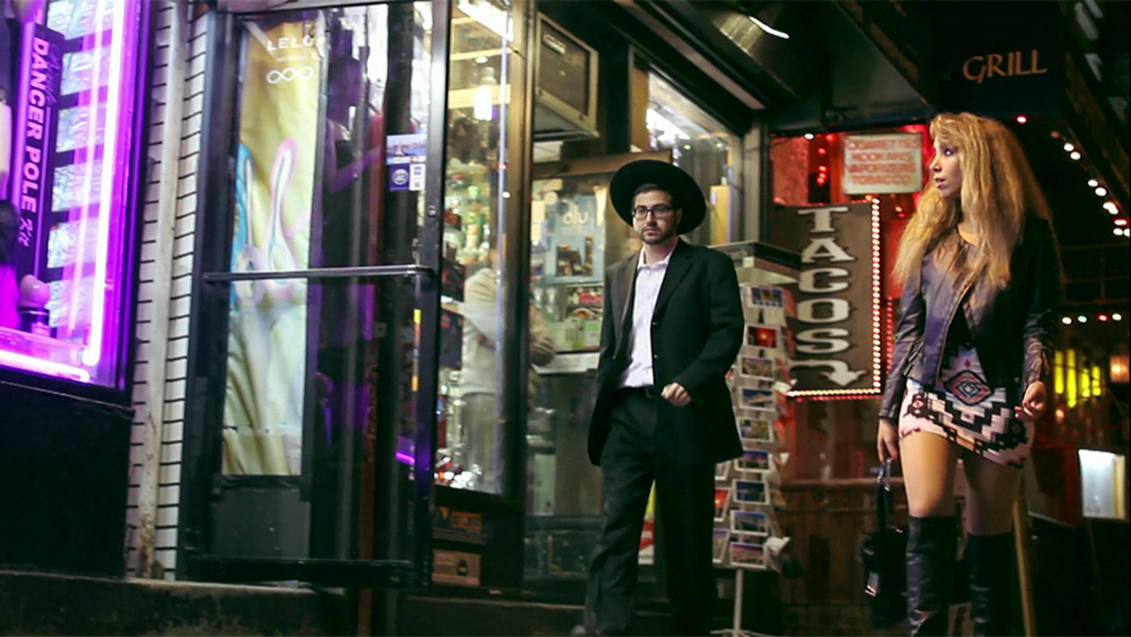 Two people walking past shops at night. The man wears a long coat and hat, the woman a leather jacket, miniskirt and boots.