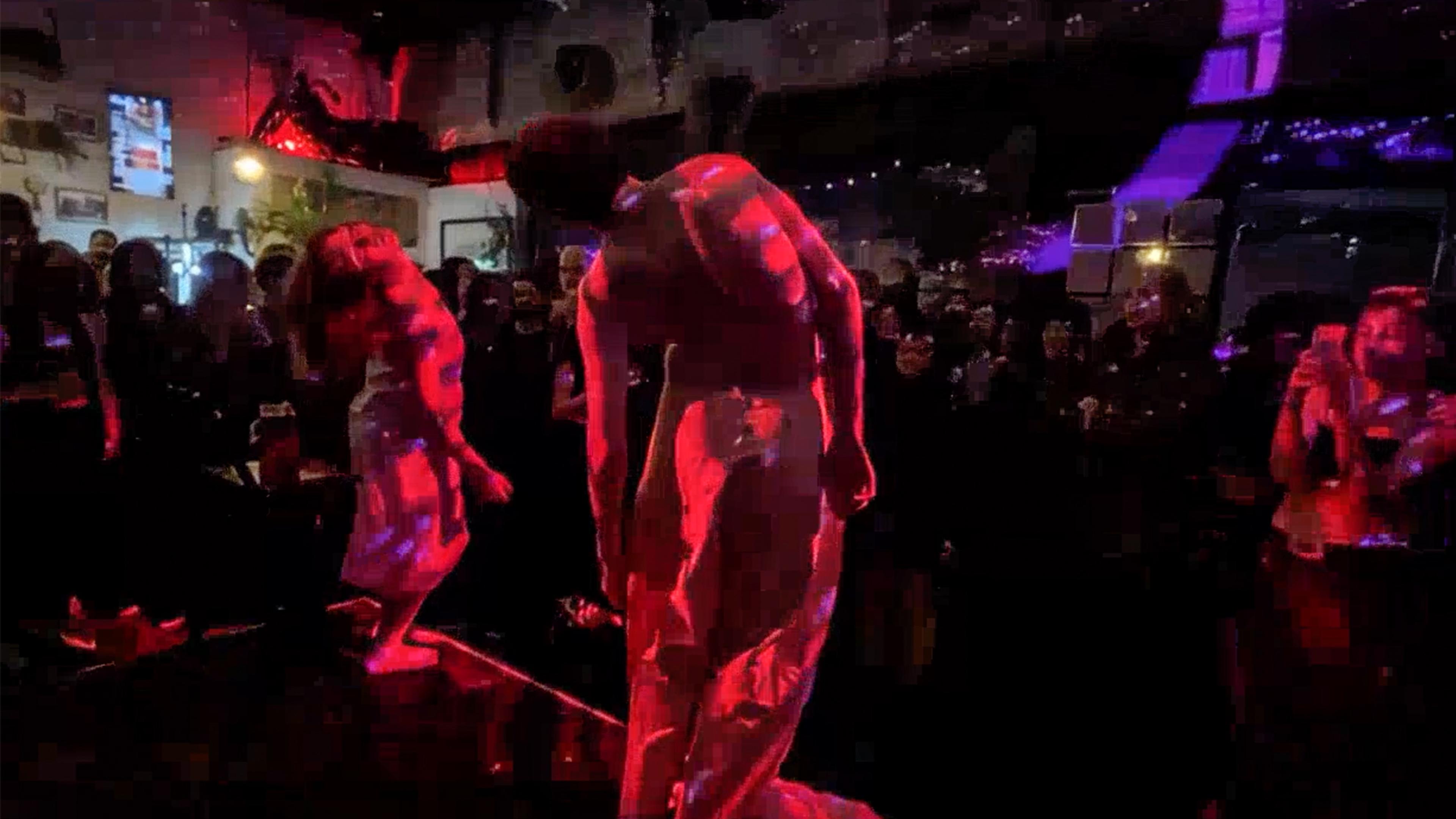 Photo of dancers in red lighting performing on stage before a crowd in a dimly lit venue.
