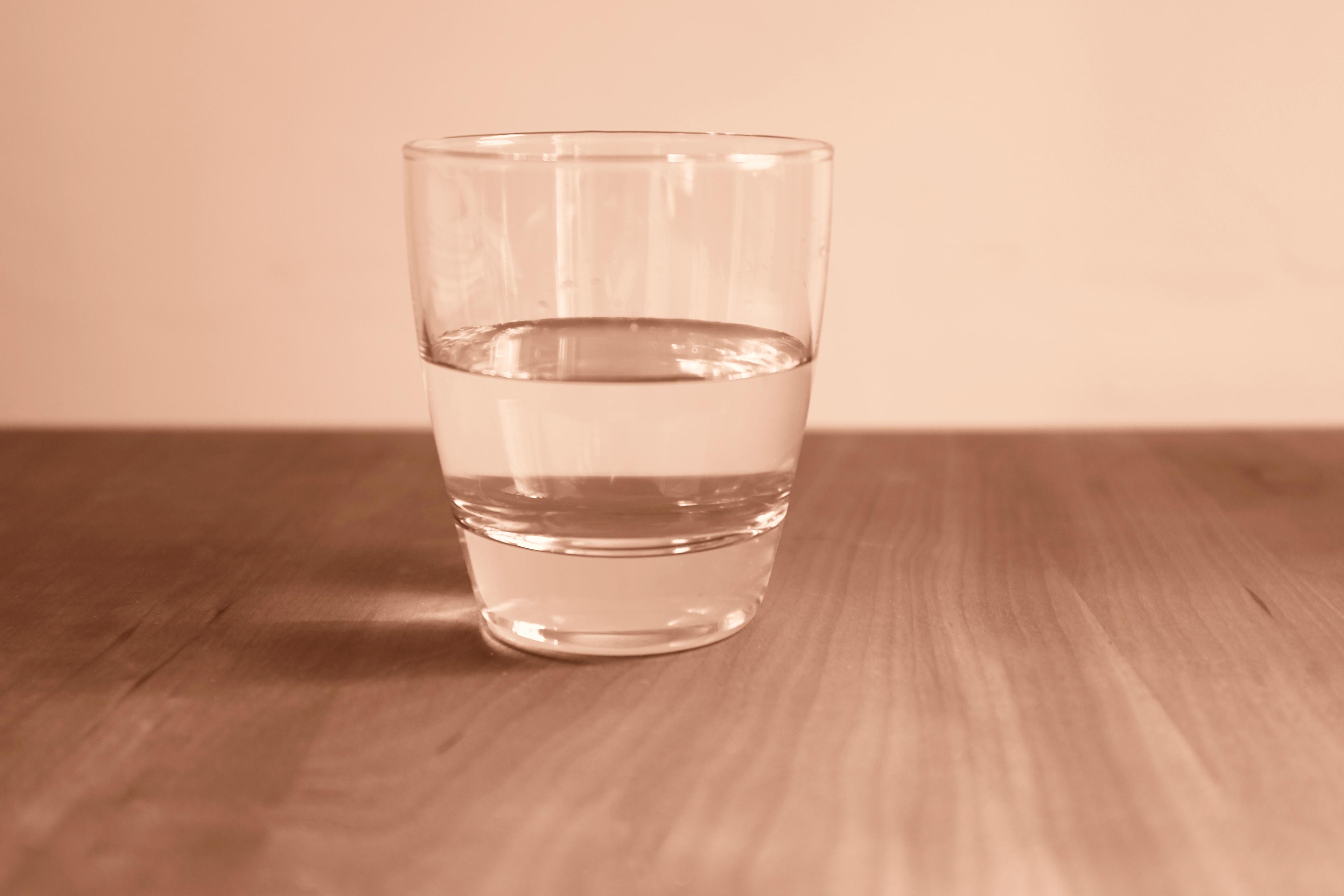 Photo of a glass half full of water on a wooden table with a beige background.