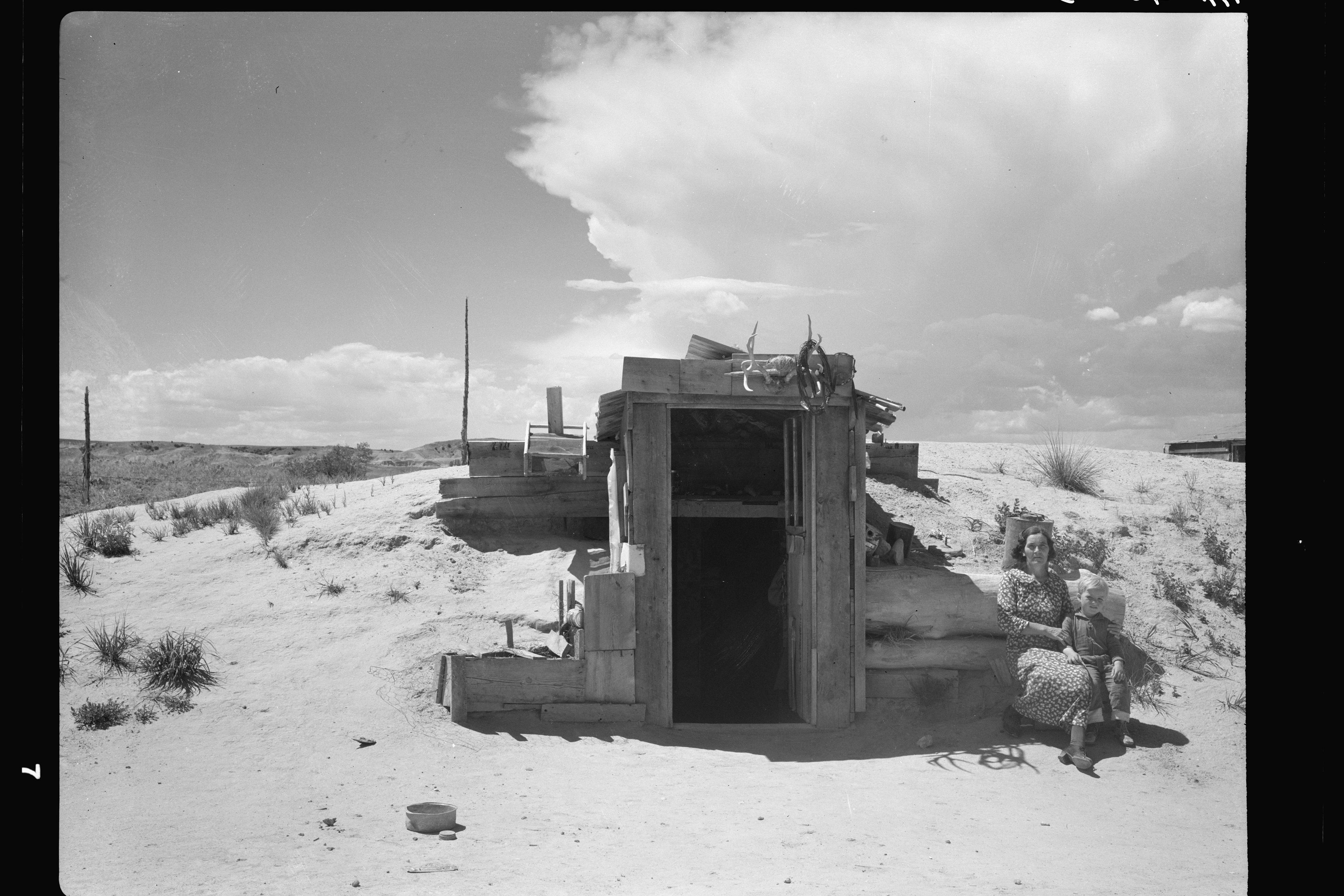 Black and white photo of a woman and child sitting outside a rustic cabin in a desert landscape under a cloudy sky.