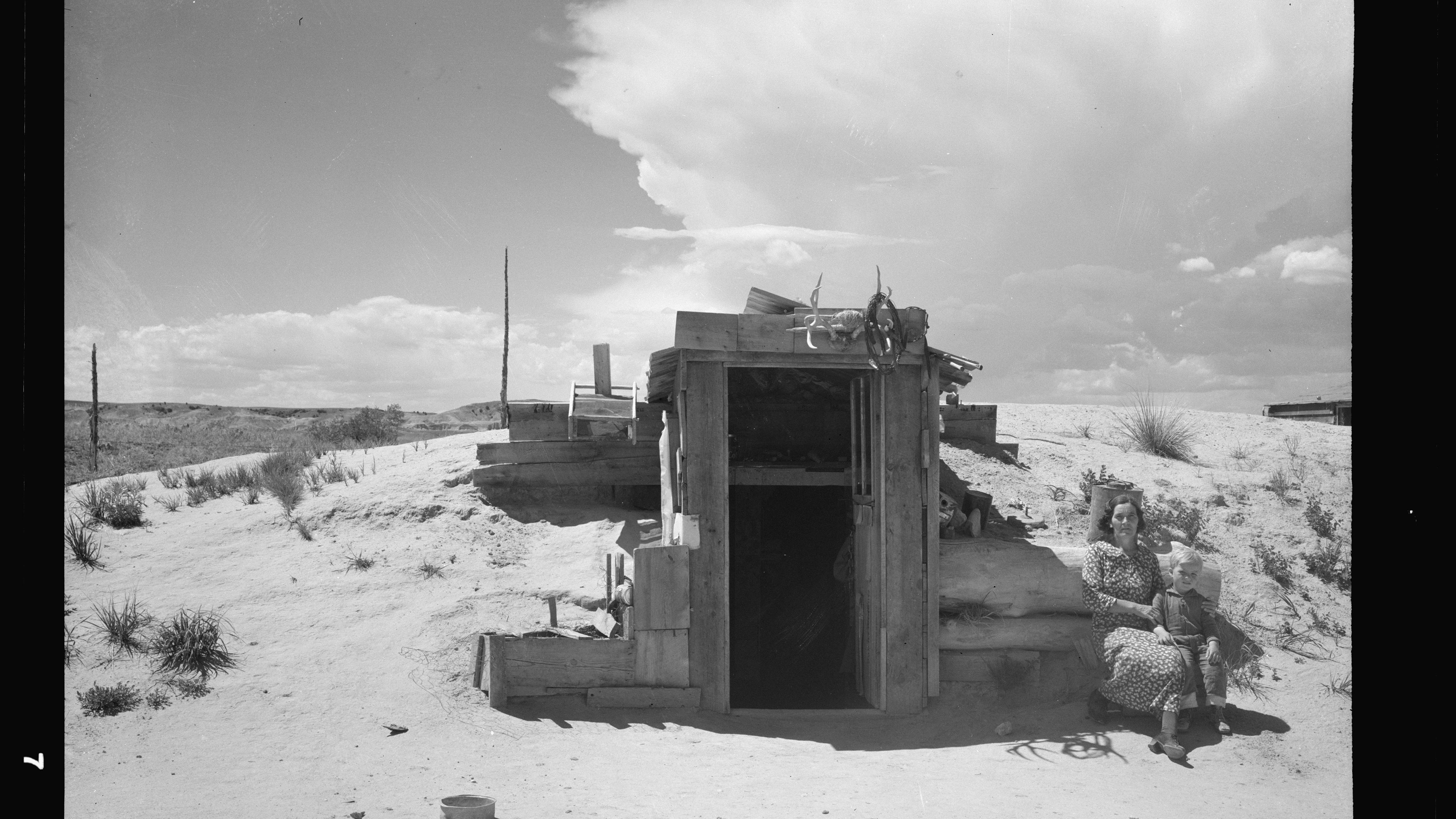 Black and white photo of a woman and child sitting outside a rustic cabin in a desert landscape under a cloudy sky.